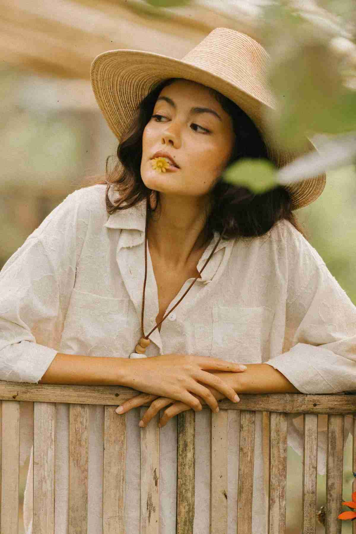 Dressed in Myrah Penaloza's Linen Nidra Button Down Set, a woman leans on a rustic wooden fence. With her straw hat and a yellow flower delicately held in her mouth, she appears contemplative amidst the blurred greenery backdrop. This limited edition ensemble, adorned with subtle florals, offers a fresh and elegant look.