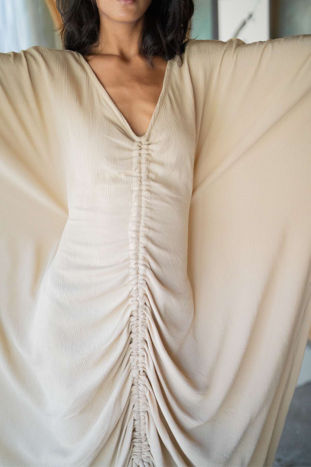 Dressed in the Virgo Moon by Myrah Penaloza, a person wearing this beige ruched dress, with its wide, flowing sleeves and v-neckline adorned with gathered details at the center, exudes ceremonial whimsy. Only the upper body is visible, yet it captures a modern and regal essence.