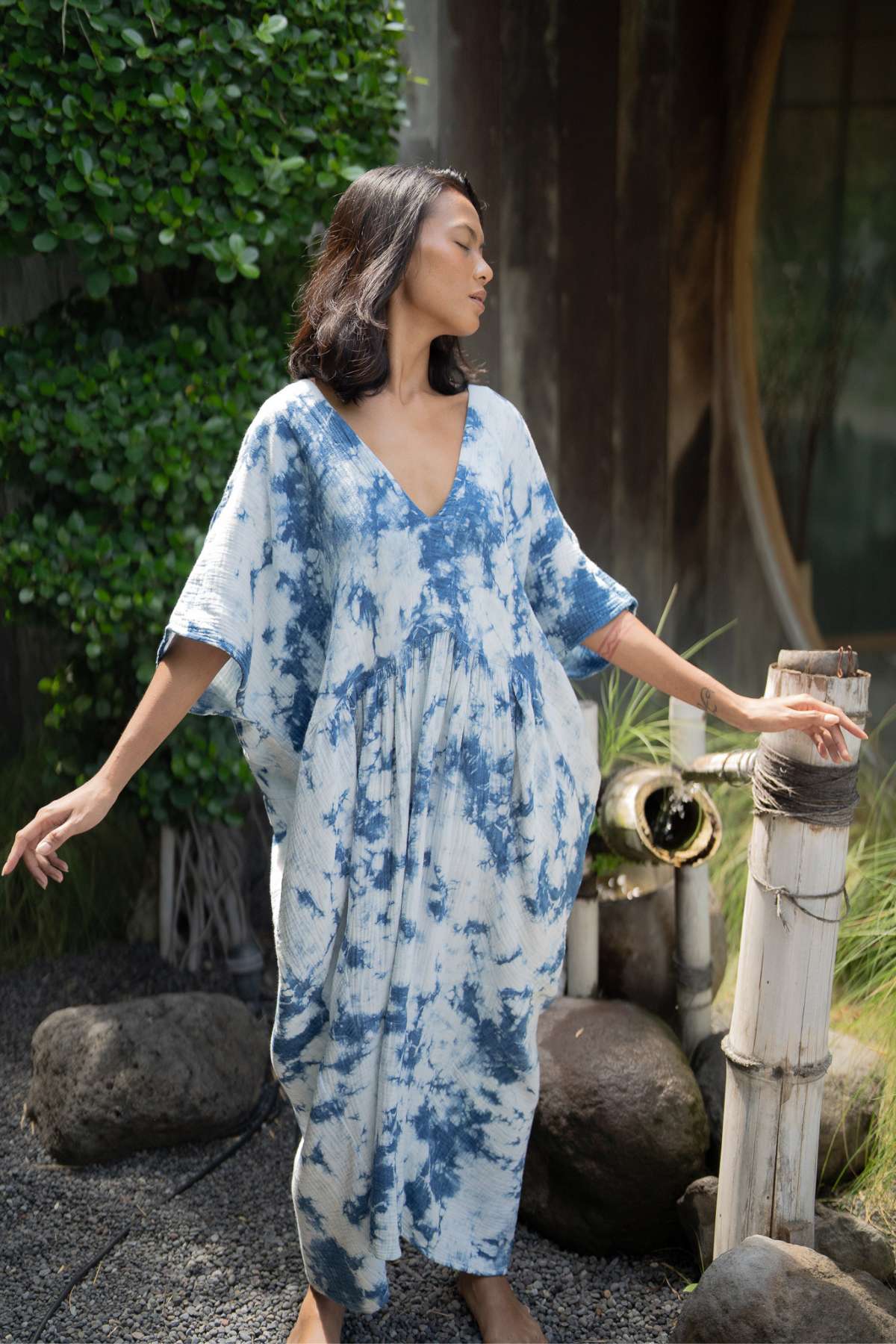 A woman wearing the Indigo Venus Gown by Myrah Penaloza stands outdoors against a backdrop of greenery and rocks. Posing gracefully with her eyes closed, she embodies effortless style, creating a peaceful and serene atmosphere.