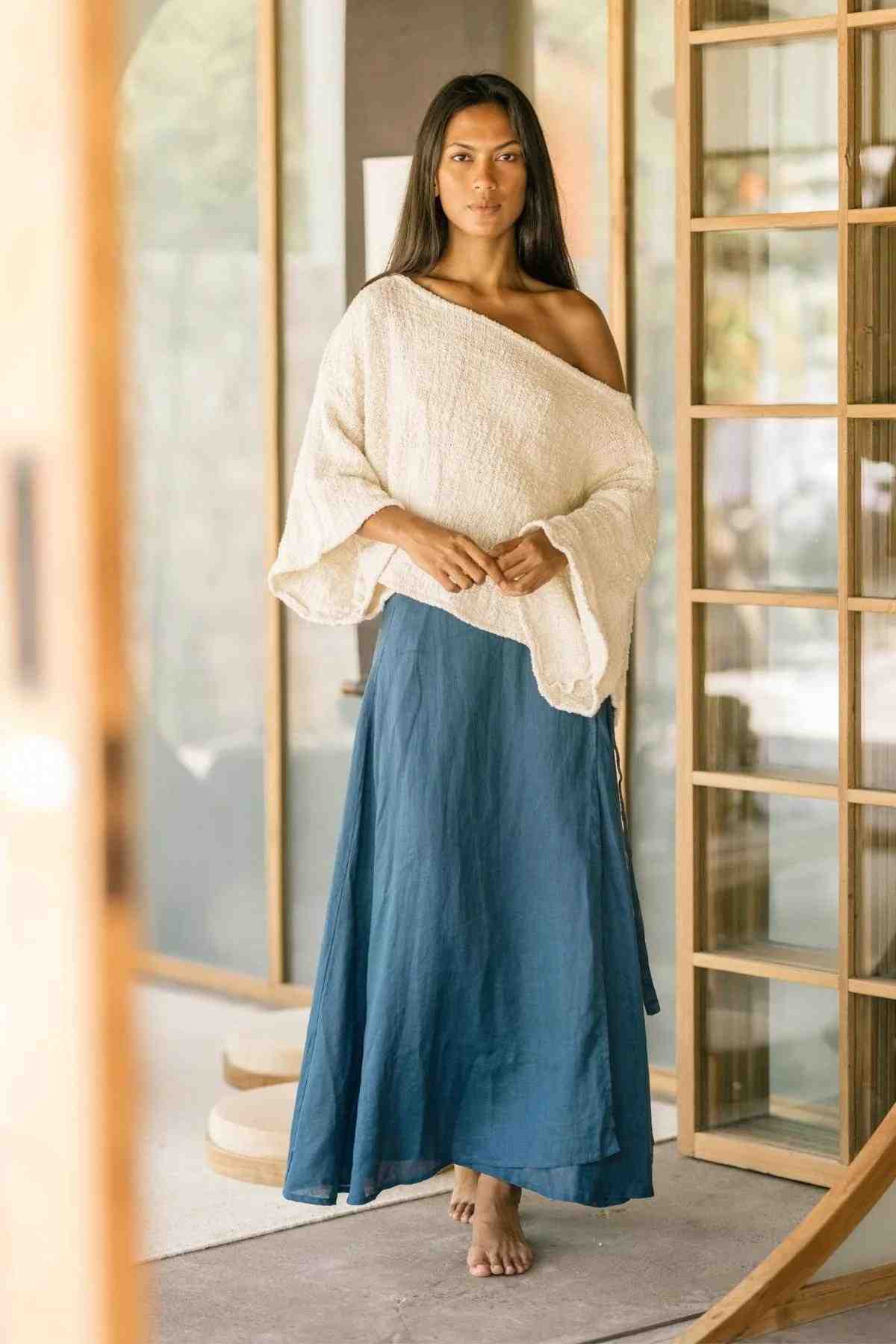 A woman stands barefoot in a well-lit, open space. She is wearing a loose, one-shouldered cream-colored top and a long, flowing Shanti Wrap Skirt made of 100% linen by Myrah Penaloza. Her straight dark hair rests on her shoulders against a background featuring wooden elements and large windows.