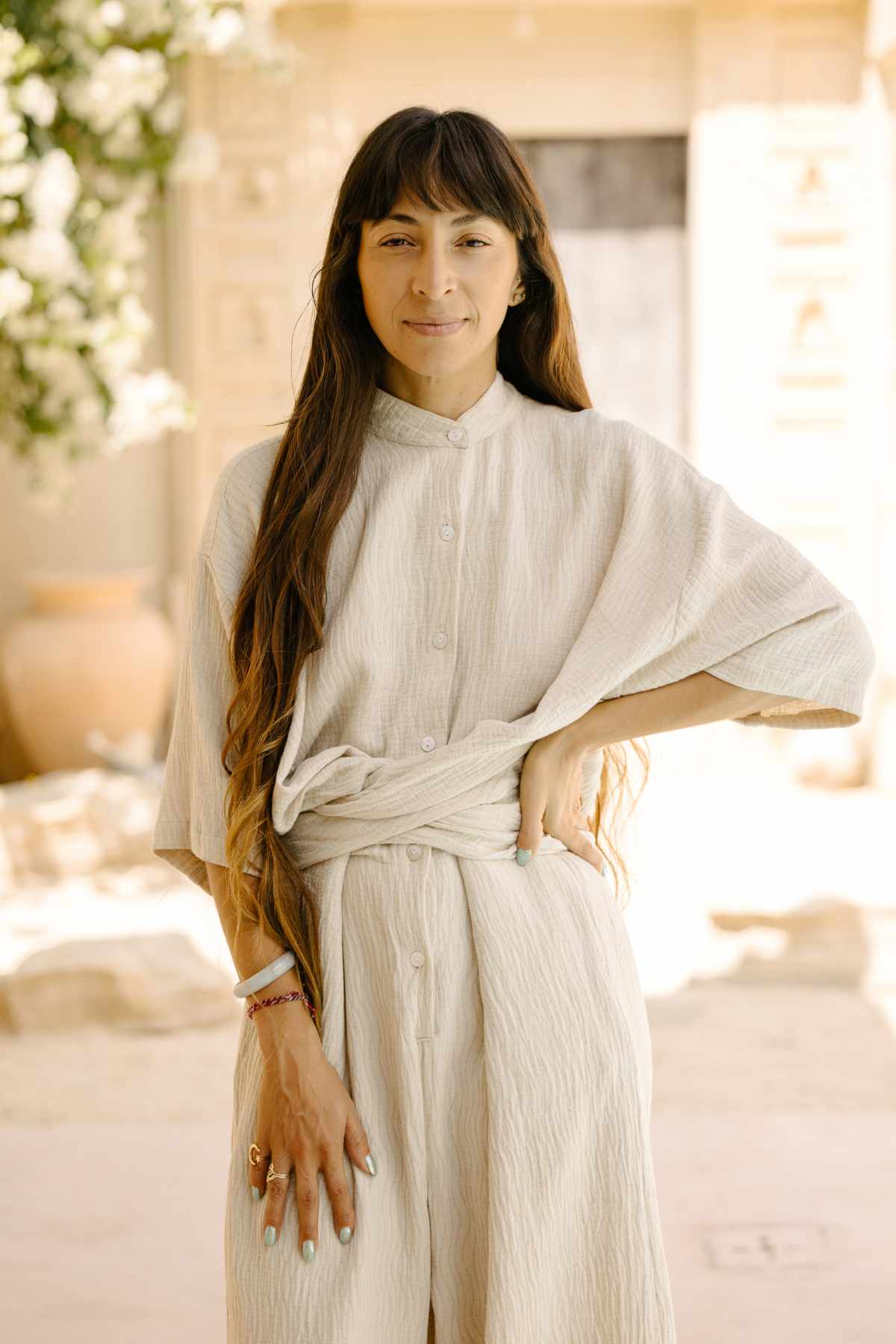 A woman with long dark hair stands confidently outside, donning the Pine Playsuit by Myrah Penaloza crafted from a textured cotton/linen blend. Her hand rests on her hip, and she has a calm expression. The background is adorned with architectural details and bathed in soft, natural lighting, enhancing her versatile wardrobe choice.