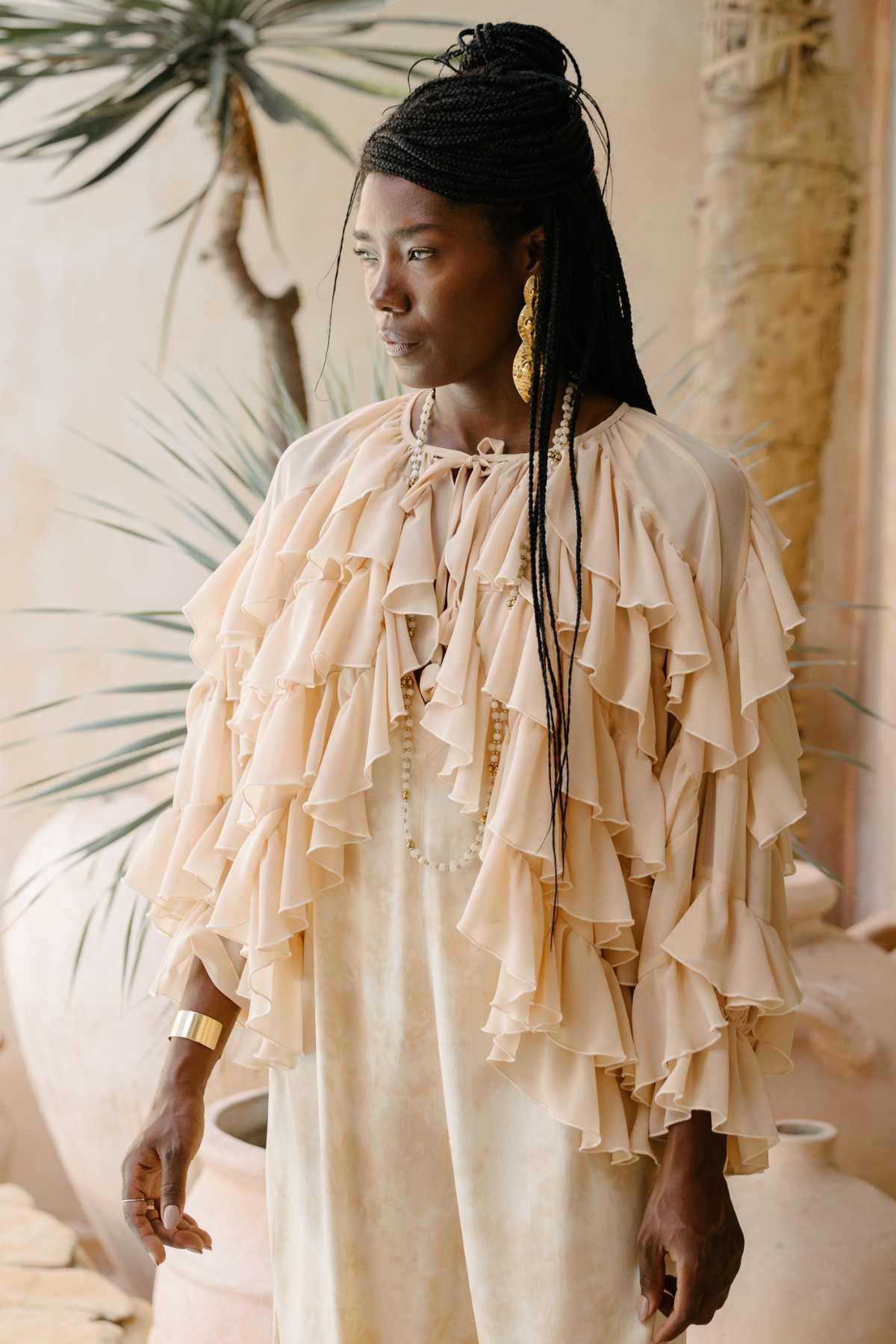 A woman stands indoors wearing the "Anusara Bloom - Made To Order" by Myrah Penaloza, a cream-colored, long-sleeved blouse with ruffles and layered flounces. Her long braided hair pairs perfectly with her gold dangling earrings and gold bracelet. Behind her, potted plants and neutral-toned decor complete the serene setting.