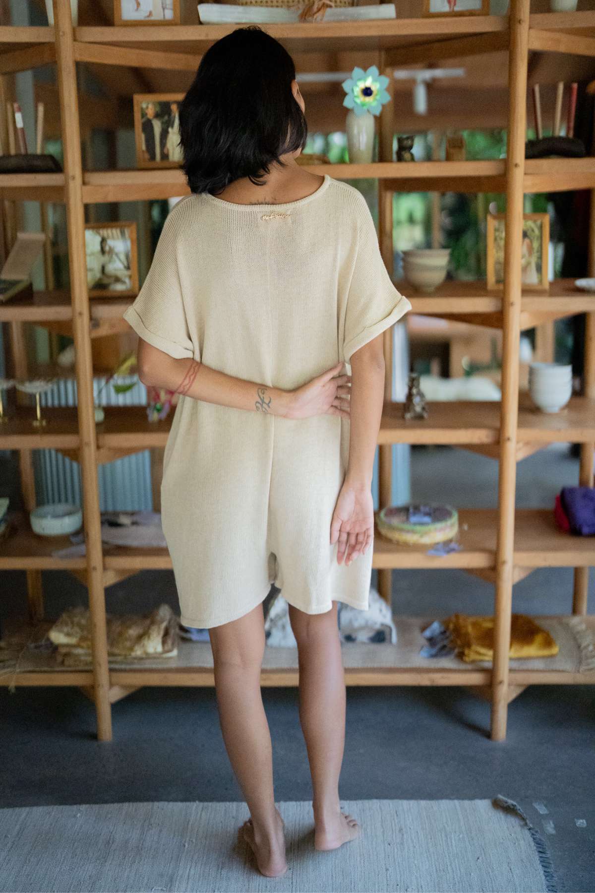 A person with dark hair stands barefoot, facing a wooden bookshelf filled with various items, wearing the Myrah Penaloza Henley Knitted Playsuit - BF, a casual cotton knit ensemble that's perfect for easy wear. Their arm rests on their lower back, surrounded by decorative objects and books in the background.