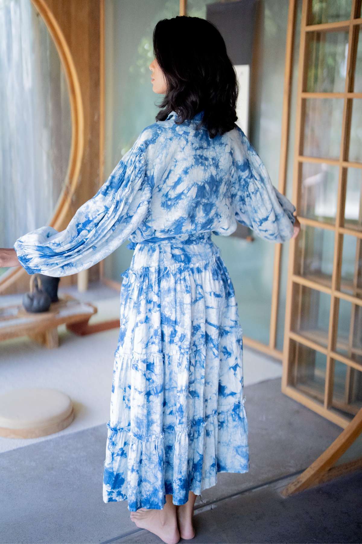 A woman in a gracefully flowing La Dama Long Sleeves Ruffles dress by Myrah Penaloza, crafted from hand tie-dyed indigo, stands barefoot on a porch. Her back is to the camera as she slightly extends her arms, embodying renaissance-inspired elegance. The setting includes wooden features and glass panels in the background.