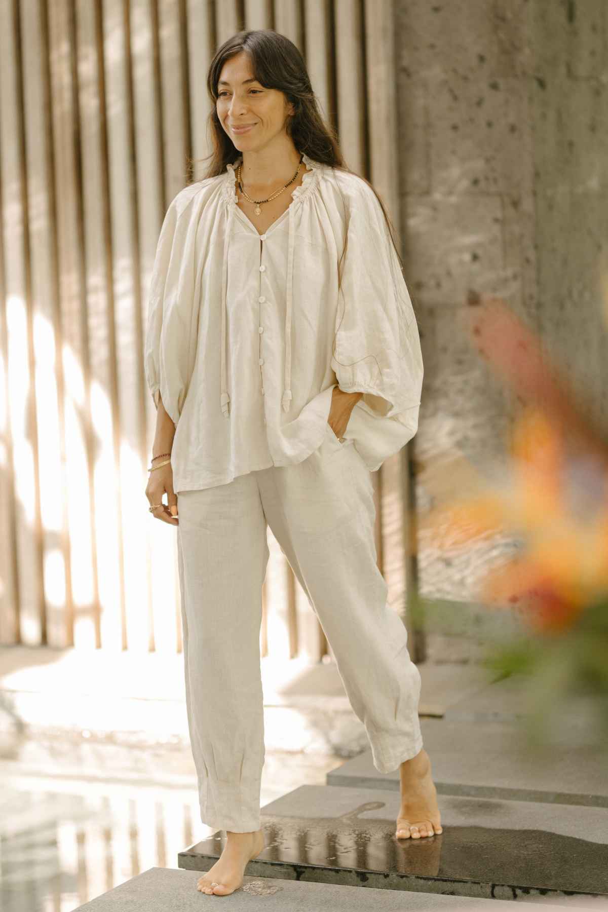 A woman with long dark hair stands barefoot in a serene, sunlit space against a wooden and stone backdrop. She is wearing a loose, cream-colored blouse paired with matching Linen Chajin Pants from Myrah Penaloza's collection, accessorized with layered necklaces and bracelets for a versatile wardrobe look.