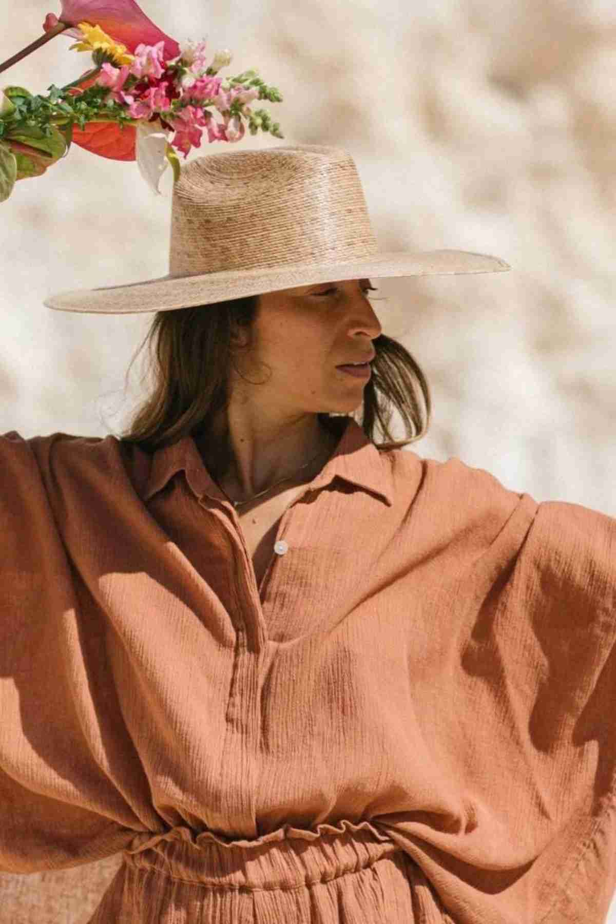 A person dressed in a Suka Button Down Top by Myrah Penaloza, designed with a loose, rust-colored style and handmade in Bali, is wearing a wide-brimmed straw hat while holding a bouquet of colorful flowers. They are outdoors against a softly blurred backdrop, embracing sustainable fashion.