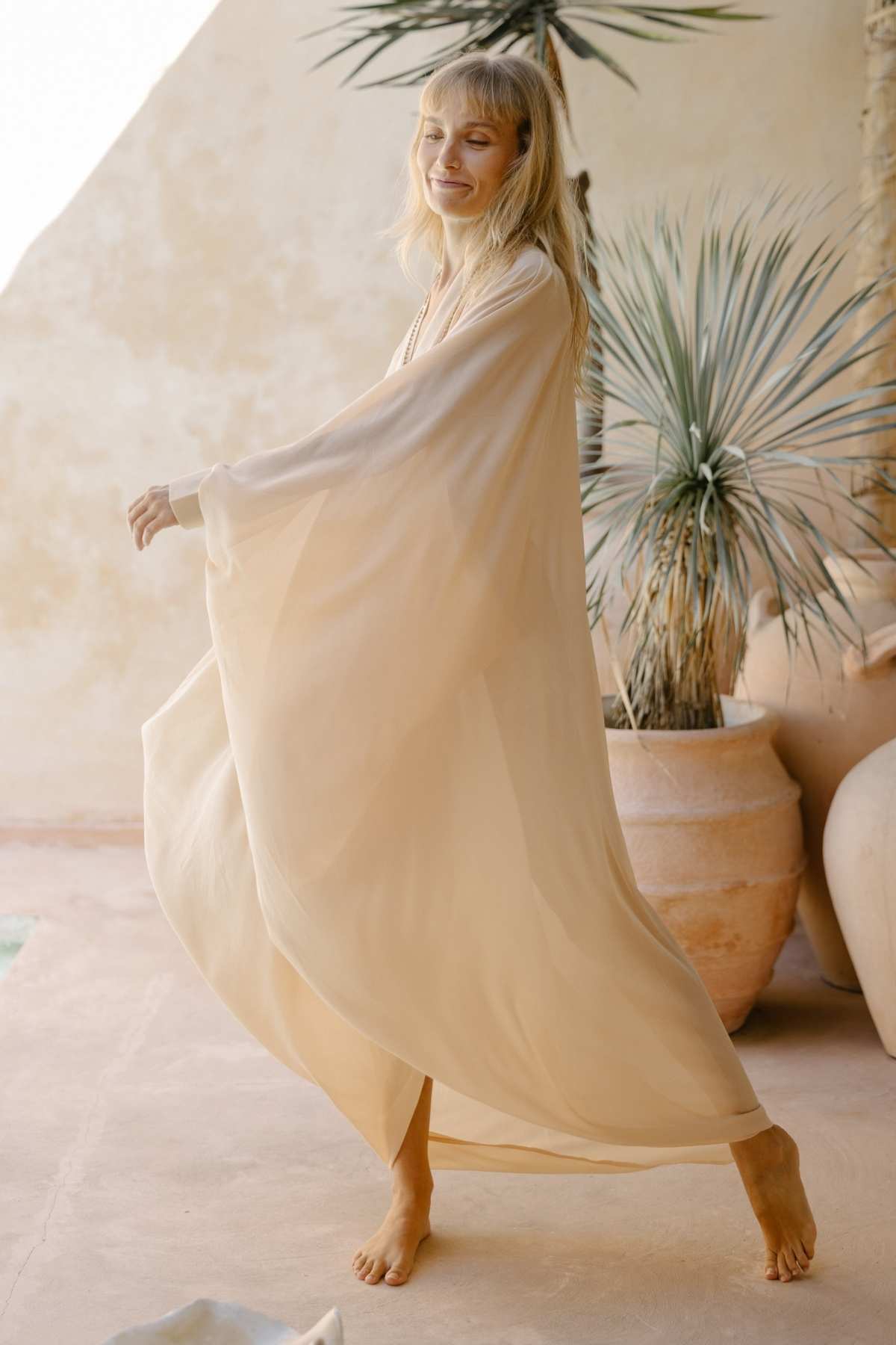 A blonde woman in a flowy Myrah Penaloza Leo Kaftan Chiffon Sheer, light beige and made of silk, gracefully walks barefoot, smiling with her eyes closed. In the background, large ceramic pots hold spiky green plants set against a light beige wall. The setting appears serene and airy, exuding luxurious power.