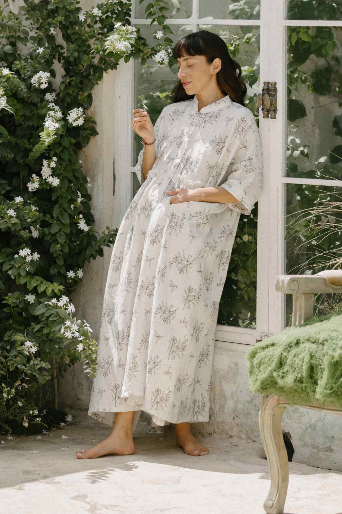 A woman stands barefoot near a large window, wearing the elegant Bona Dress from the Cha Dao Tea Collection by Myrah Penaloza, made of 100% linen with a tea leaf eco print. She leans against the wall, surrounded by lush, green foliage. To her right is a small, cushioned seat adorned with a green throw.