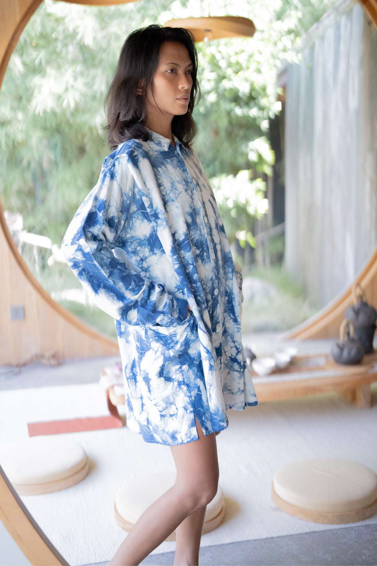 Against a backdrop of minimalist decor and natural light streaming through lush greenery outside, an individual stands indoors in front of a circular window, wearing the Myrah Penaloza Tender Jacket, an oversized garment hand-dyed in indigo. This eclectic piece adds depth to the serene scene.