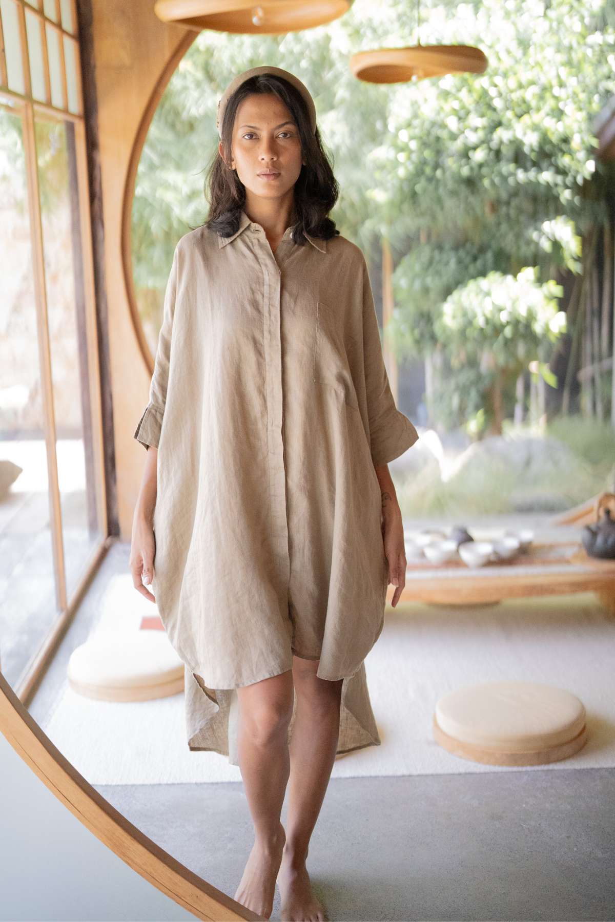 A person stands indoors, highlighting the versatile styling of a loose, beige Linen Suka Button Down Long Top - BF by Myrah Penaloza. The room is enhanced with round windows and natural elements, offering views of lush greenery outside. This serene, minimalist design is completed with cushions and a wooden floor.