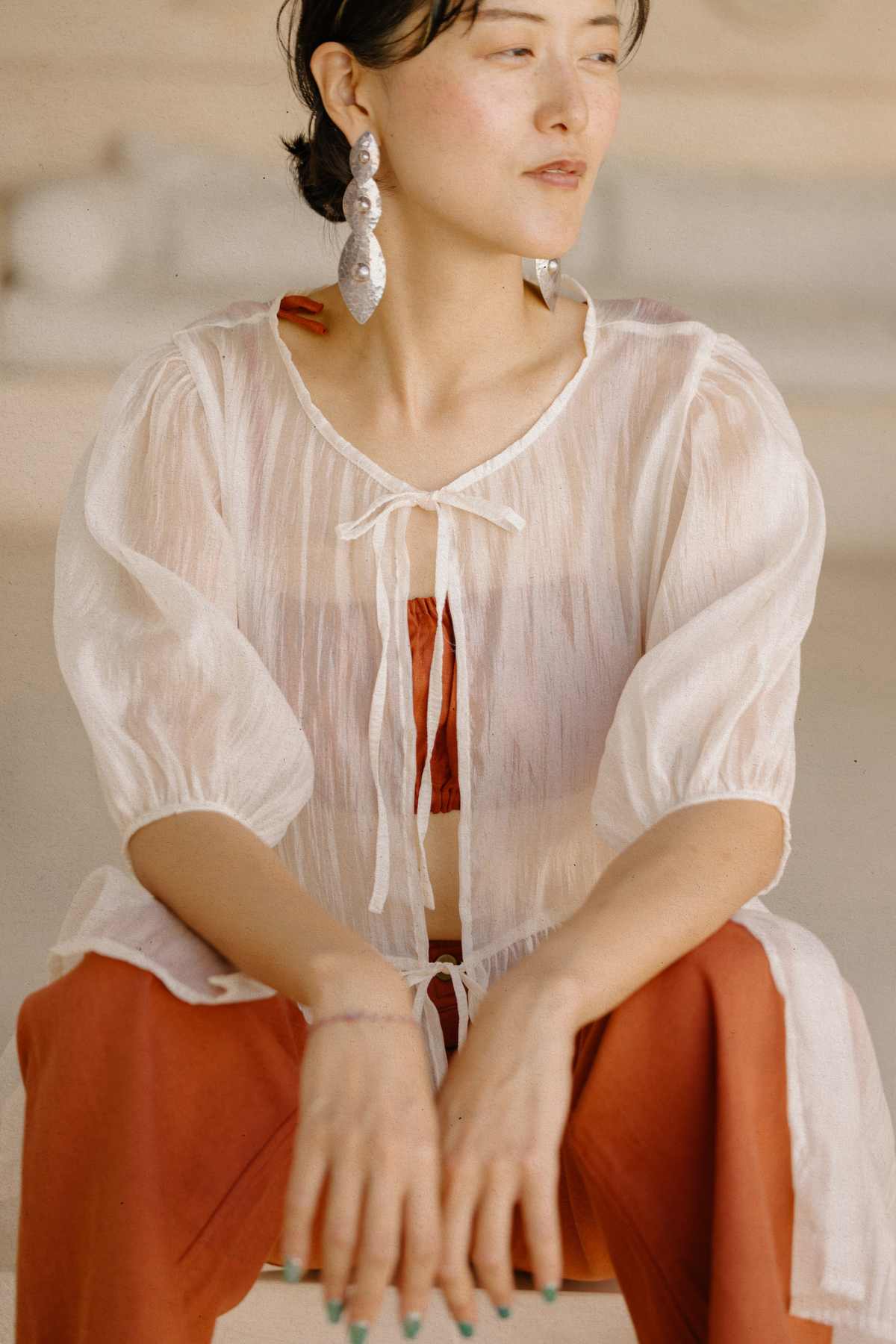 A person with short dark hair is seated, wearing the Myrah Penaloza Dynasty Sheer Silk Dress layered over an orange outfit. The dress features puffy sleeves and long, dangling earrings. They look to the side with a peaceful expression, showcasing this layering essential.