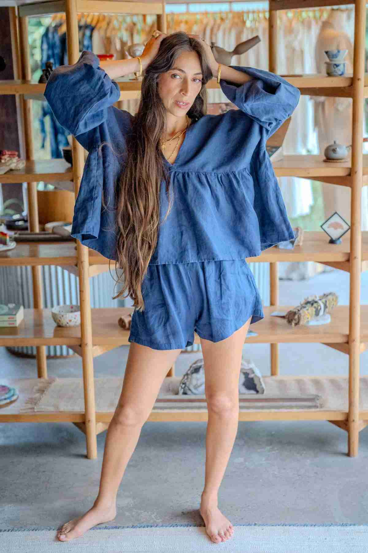 A woman with long brown hair wearing a blue, loose-fitting Eva Linen Top by Myrah Penaloza stands barefoot in front of a wooden shelf. She is holding her hair with one hand and looking into the camera. The shelf behind her displays clothes, books, and decorative items.