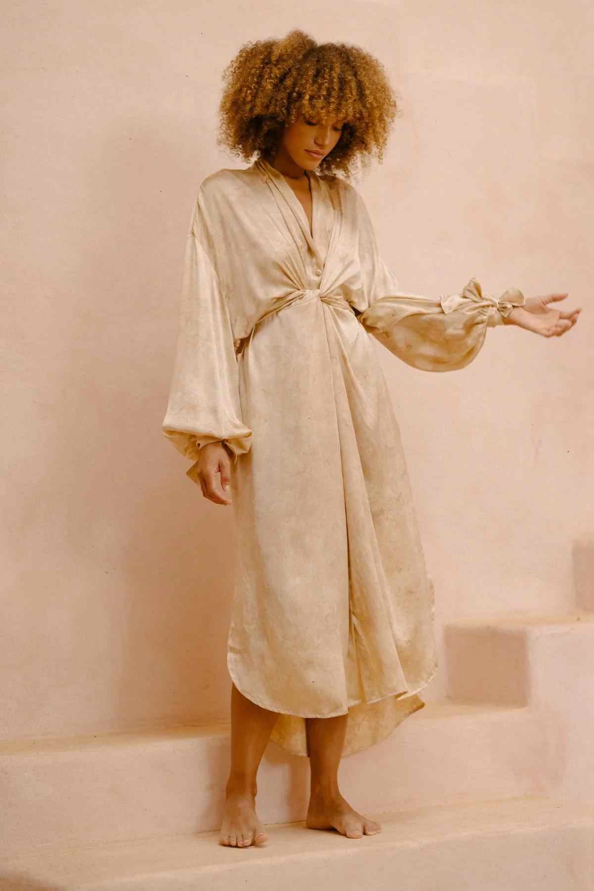 A person with curly hair stands on stairs, wearing a long, flowing beige Silk Inanna Wrap Gown by Myrah Penaloza. The gown features billowy sleeves and is made from botanically hand-dyed silk. Their gaze is downward, with one arm extended against a neutral, earthy-toned wall backdrop.