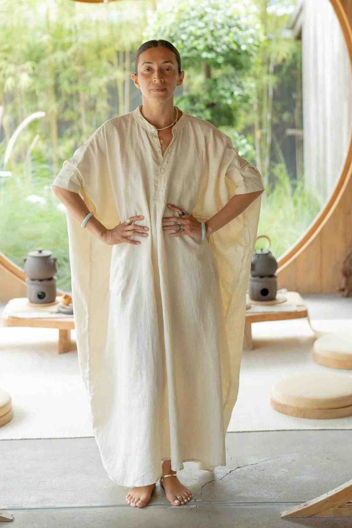 A person stands confidently indoors, adorned in the elegant La Majia Kaftan by Myrah Penaloza—a long, cream-colored, flowing linen dress with short sleeves. They have their hands on their hips. Behind them is a circular wooden opening that appears to lead to a garden outside. The setting is minimalistic with round cushions, evoking a Frida Kahlo vibe.