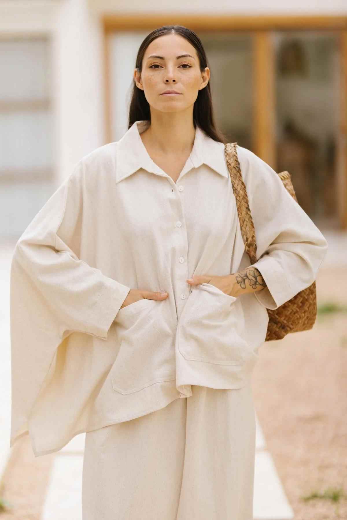 A person with long, dark hair stands facing the camera, wearing the Myrah Penaloza Grace Jacket & Mona Pant Set in Cotton Linen Natural. They have their hands in their pockets and are holding a woven bag on their shoulder. The background is out-of-focus but appears to be outdoors.