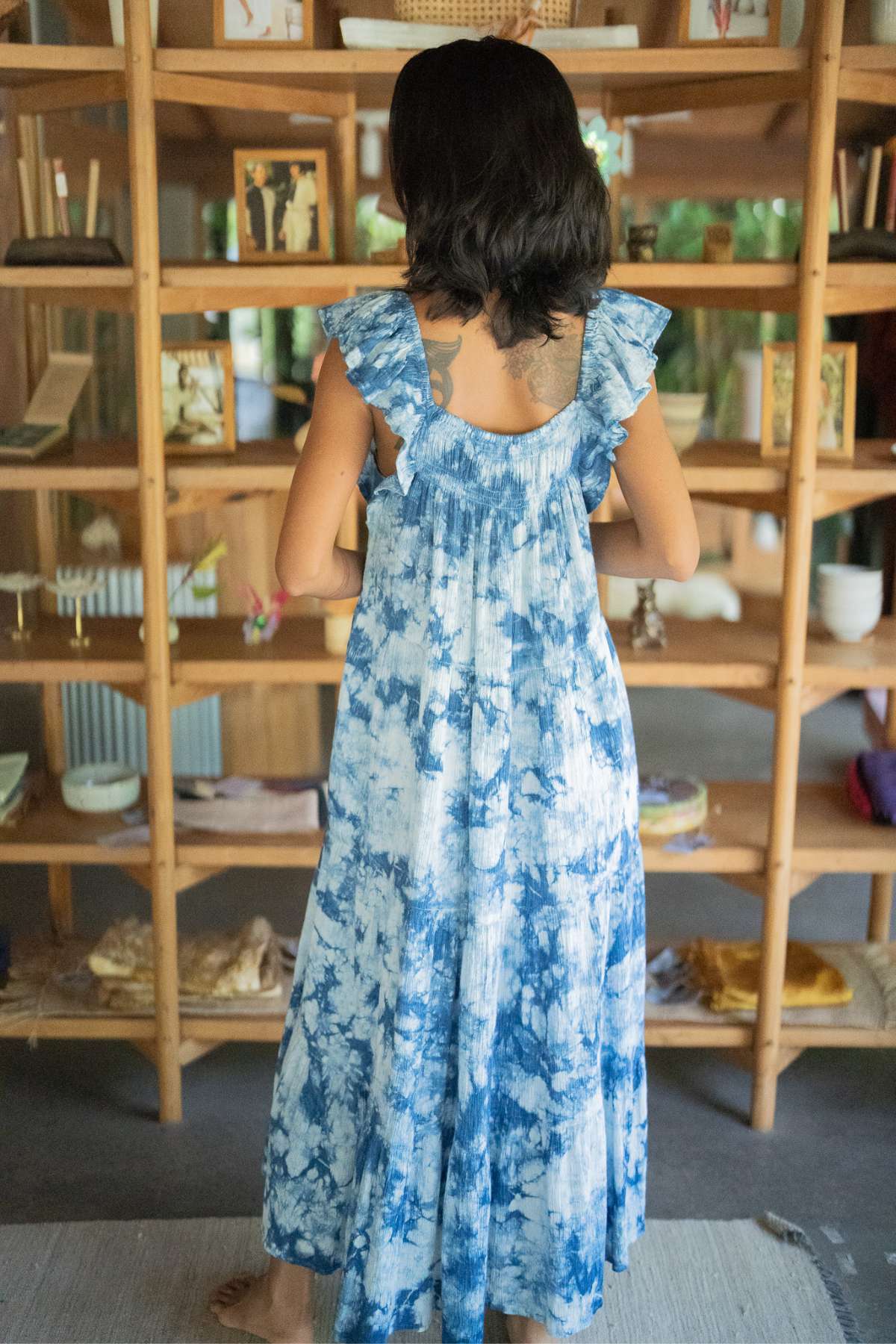 A woman with shoulder-length dark hair stands with her back to the camera, wearing the Indigo Dreams Anna Yemaya Gown by Myrah Penaloza. She is in front of a wooden bookshelf filled with various items and framed pictures.