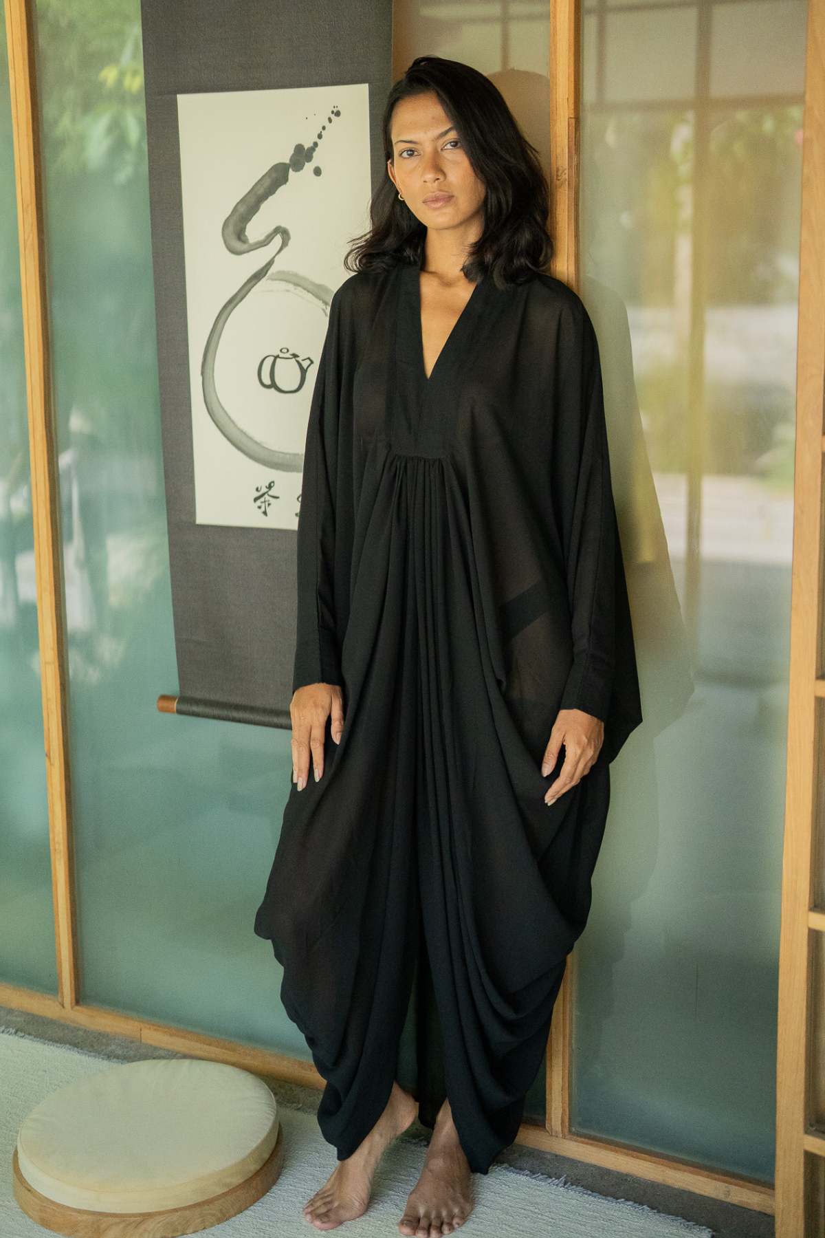 A woman stands barefoot on a light-colored floor, elegantly draped in a luxurious Leo Kaftan Chiffon Sheer by Myrah Penaloza. She is positioned before a traditional wall adorned with a hanging scroll of calligraphy, as sunlight softly filters through the window behind her.