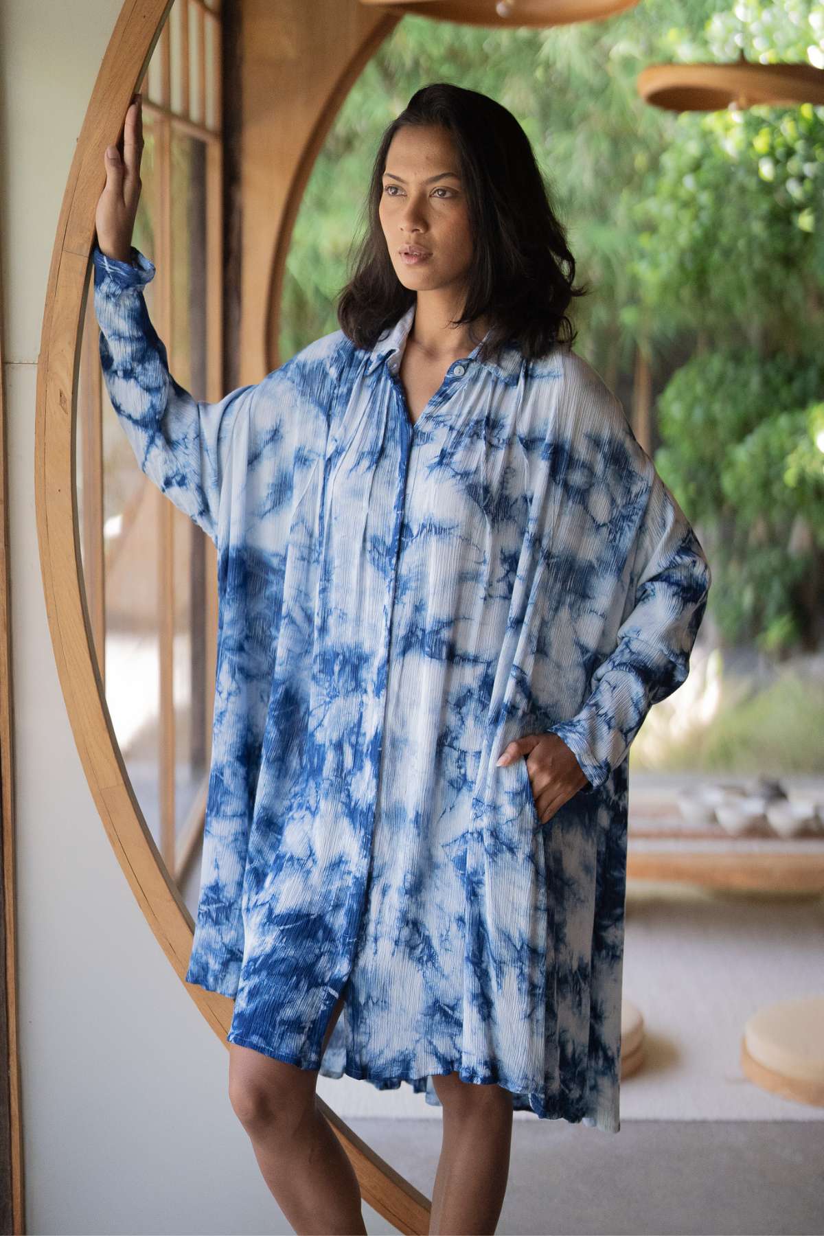 A woman stands by a circular window, adorned in Myrah Penaloza's Indigo Dreams Kundalini Gown Short, featuring a blue and white tie-dye pattern. She gracefully places her left hand on the frame while her right hand rests in her pocket. Sunlight streams through, illuminating the vibrant greenery outside.