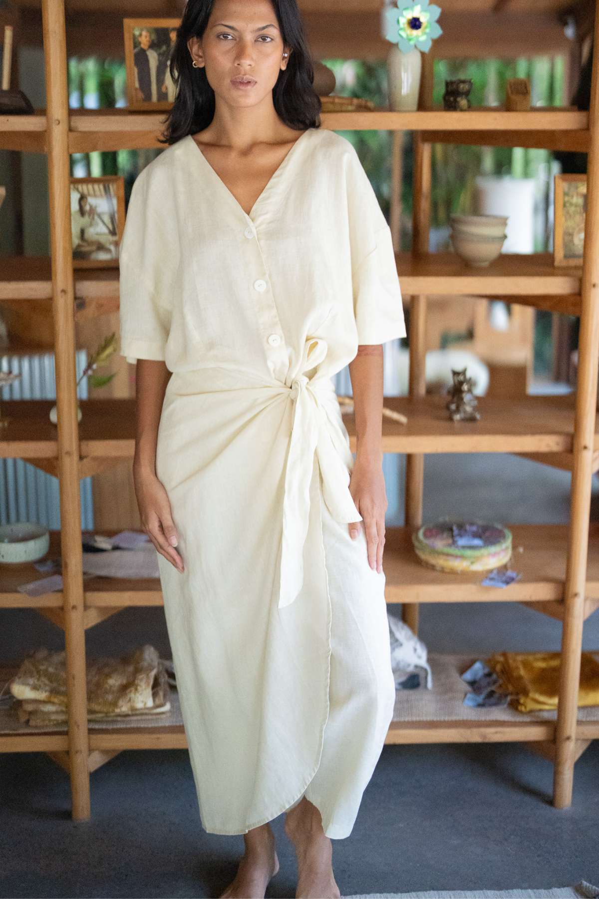 A person stands barefoot indoors, adorned in the Temple Wrap Playsuit Linen (PR) by Myrah Penaloza, which features a light-colored button-up design with an oversized fit and tied waist, reminiscent of a sarong. They are positioned in front of a wooden shelf filled with various decorative items like vases and small artworks that evoke Balinese culture.