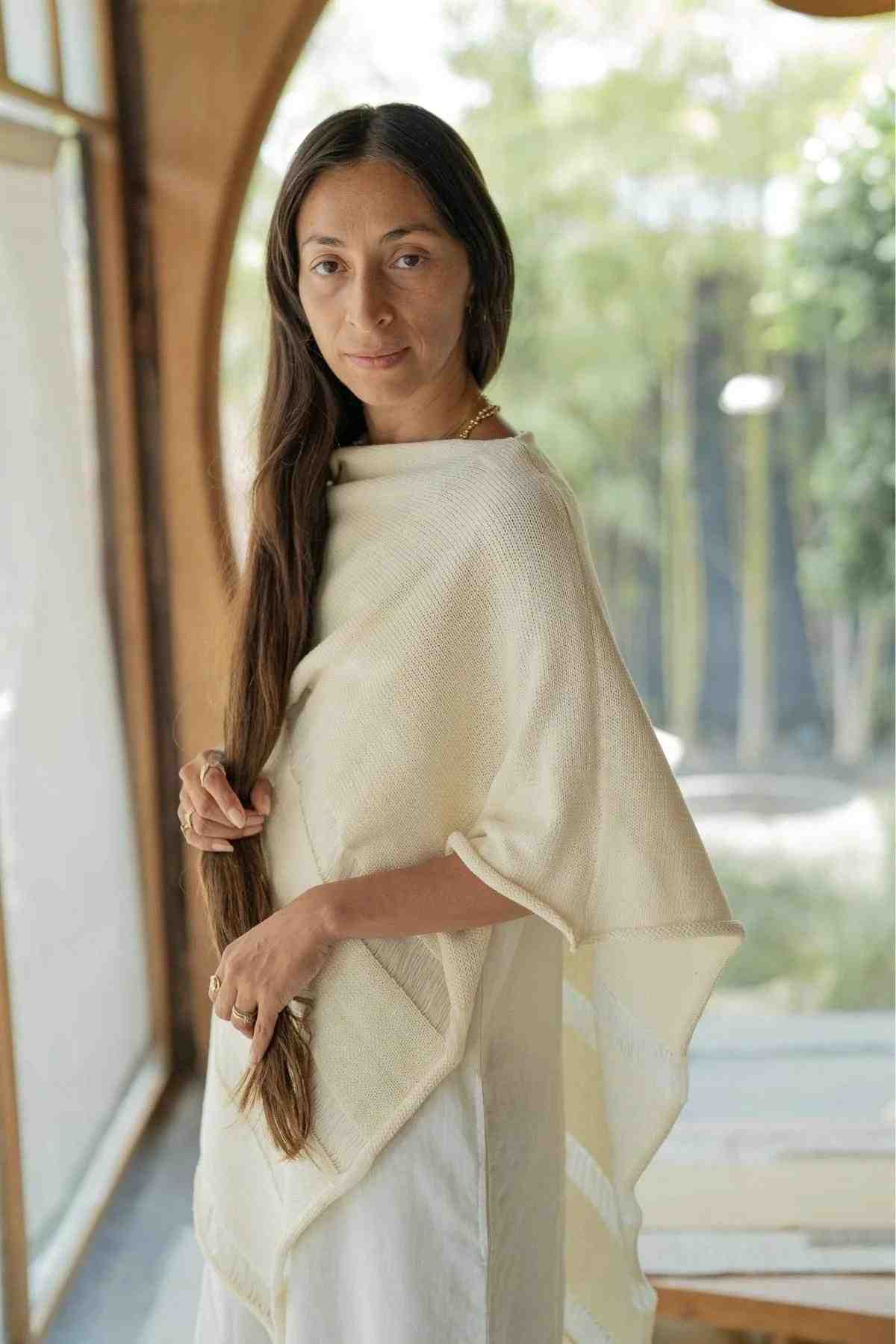 A person with long, dark hair is standing indoors near a large window. They are wearing an effortlessly chic Frida Poncho by Myrah Penaloza over a white outfit and looking directly at the camera. The background is bright with greenery visible through the window.