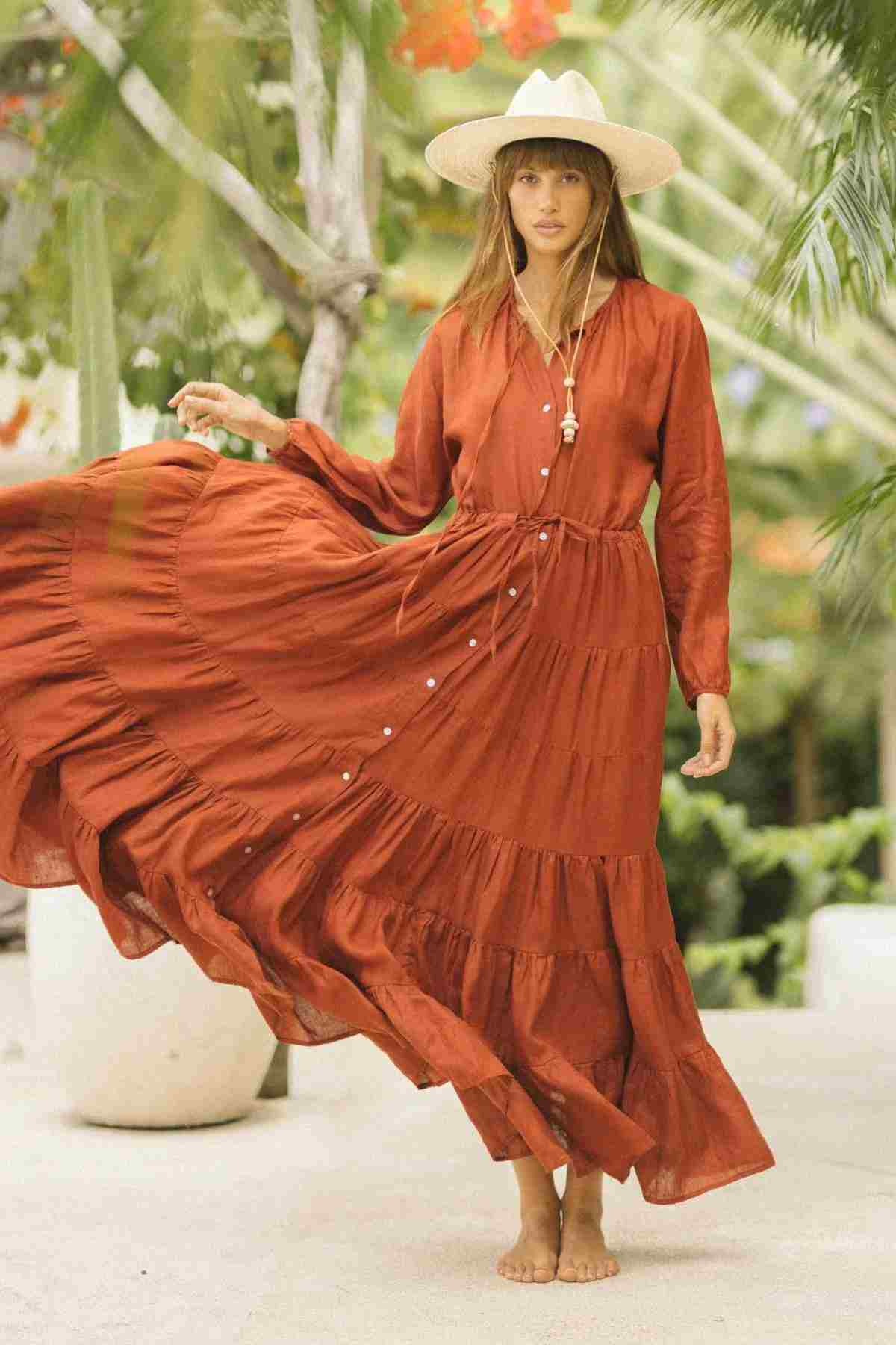 A woman stands barefoot in a lush garden, adorned in the La Mexicana Gown by Myrah Penaloza, showcasing its rust-colored elegance with an adjustable waist. She elegantly lifts one side to reveal the gown’s tiered design while wearing a wide-brimmed hat. The scene is set against a backdrop of palm leaves and large potted plants.