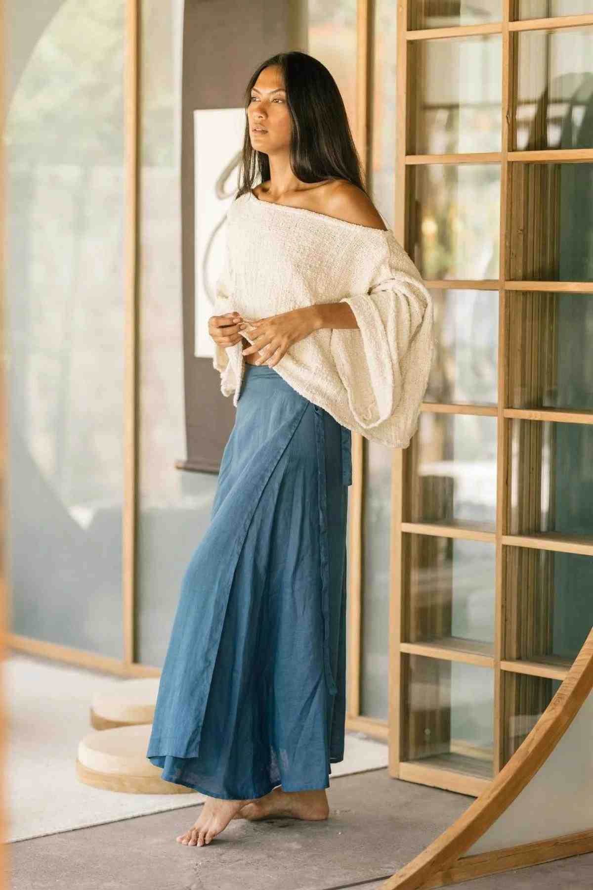 A woman with long, dark hair stands barefoot on a porch, wearing a loose off-the-shoulder white sweater and an adjustable Shanti Wrap Skirt by Myrah Penaloza, crafted from 100% linen. She looks thoughtful as she gazes into the distance, with her hands lightly clasped in front of her.