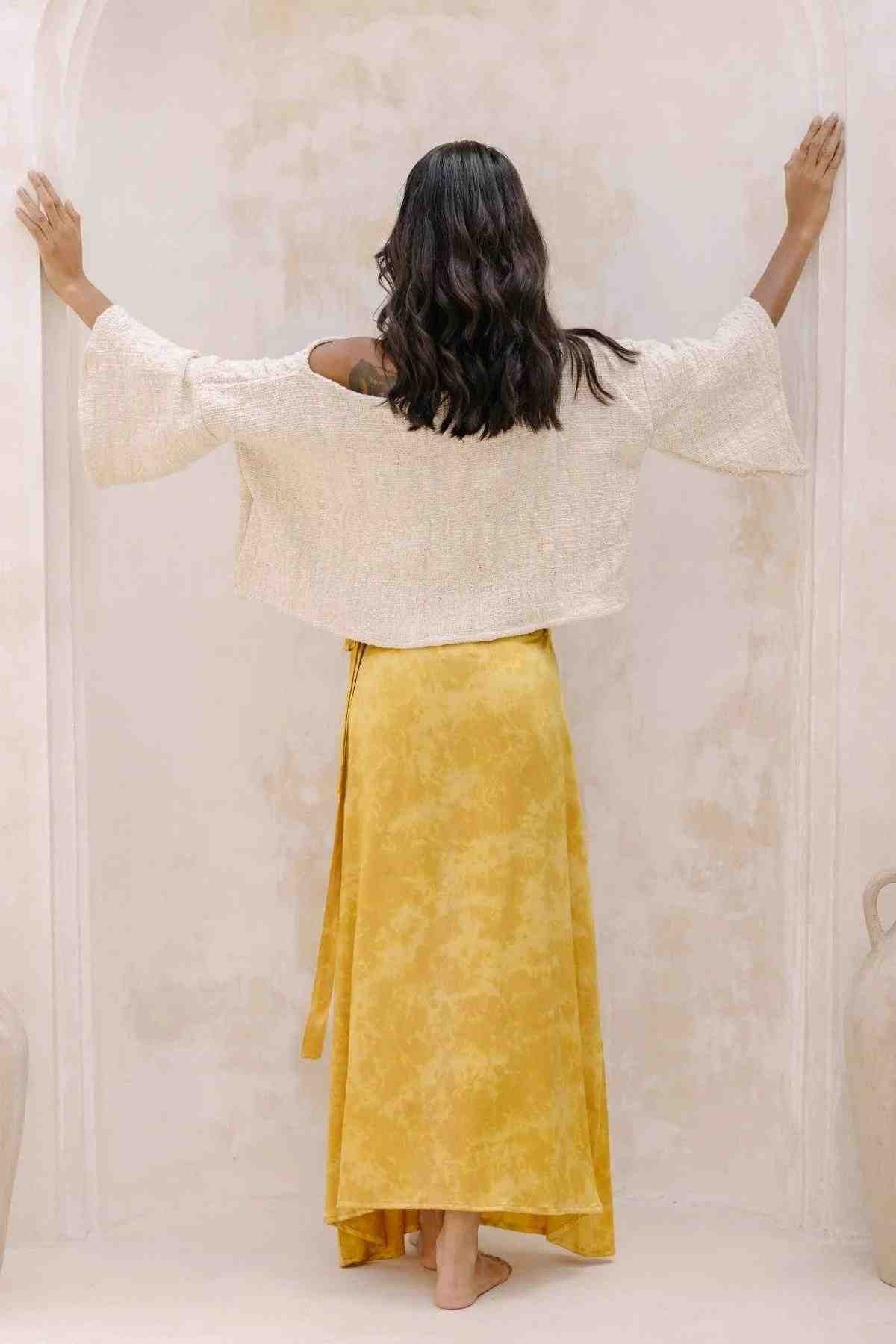 A person with long dark hair stands barefoot with their back to the camera, wearing the Spirit Top and Shanti Wrap Skirt Set by Myrah Penaloza. They are dressed in a white, long-sleeved top paired with an adjustable yellow maxi skirt made from 100% linen. Their arms are extended, touching the wall on either side in what appears to be a neutral-hued, minimalist room.