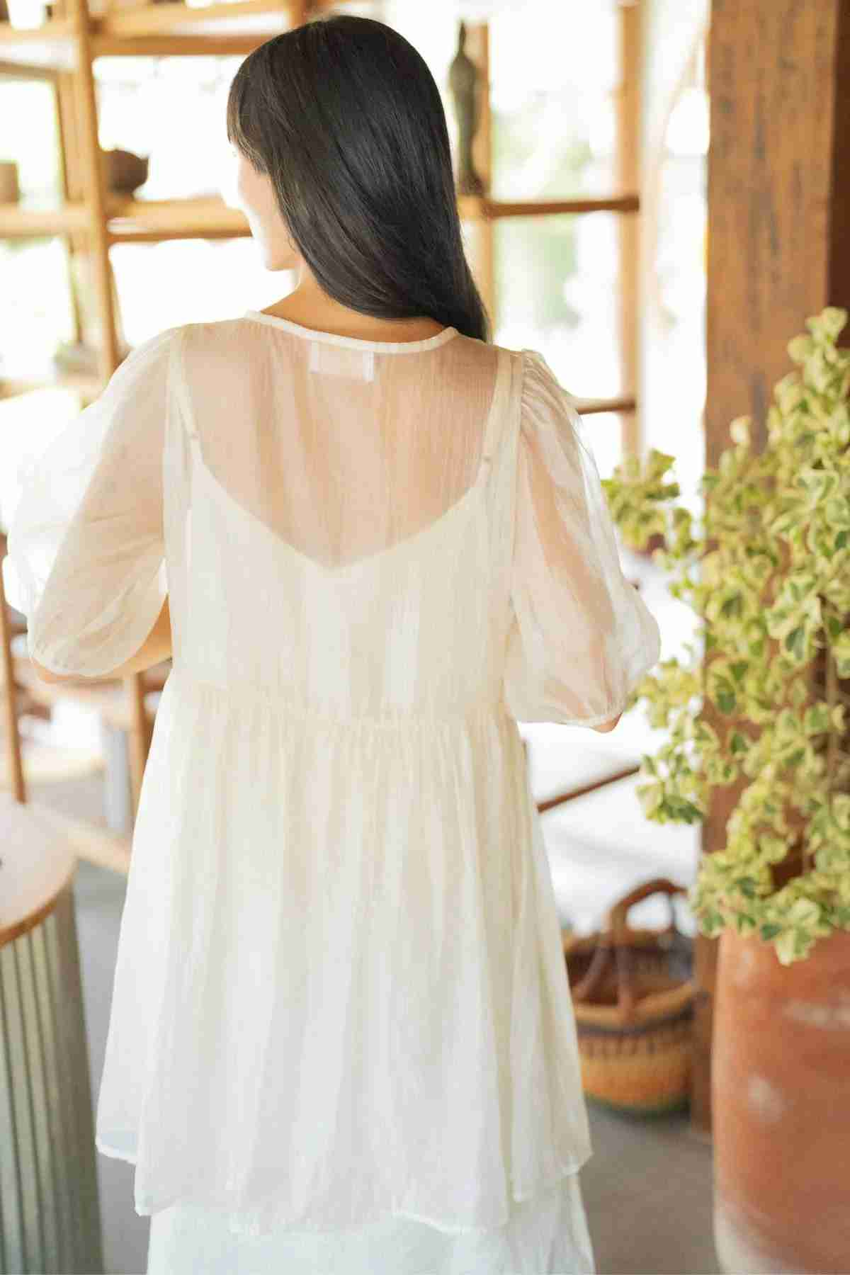 A woman with long dark hair is facing away, adorned in the Dynasty Sheer Silk Dress by Myrah Penaloza, a light and delicate silk chiffon garment. She stands in a well-lit room with wooden shelves and plants, creating a cozy and serene atmosphere.