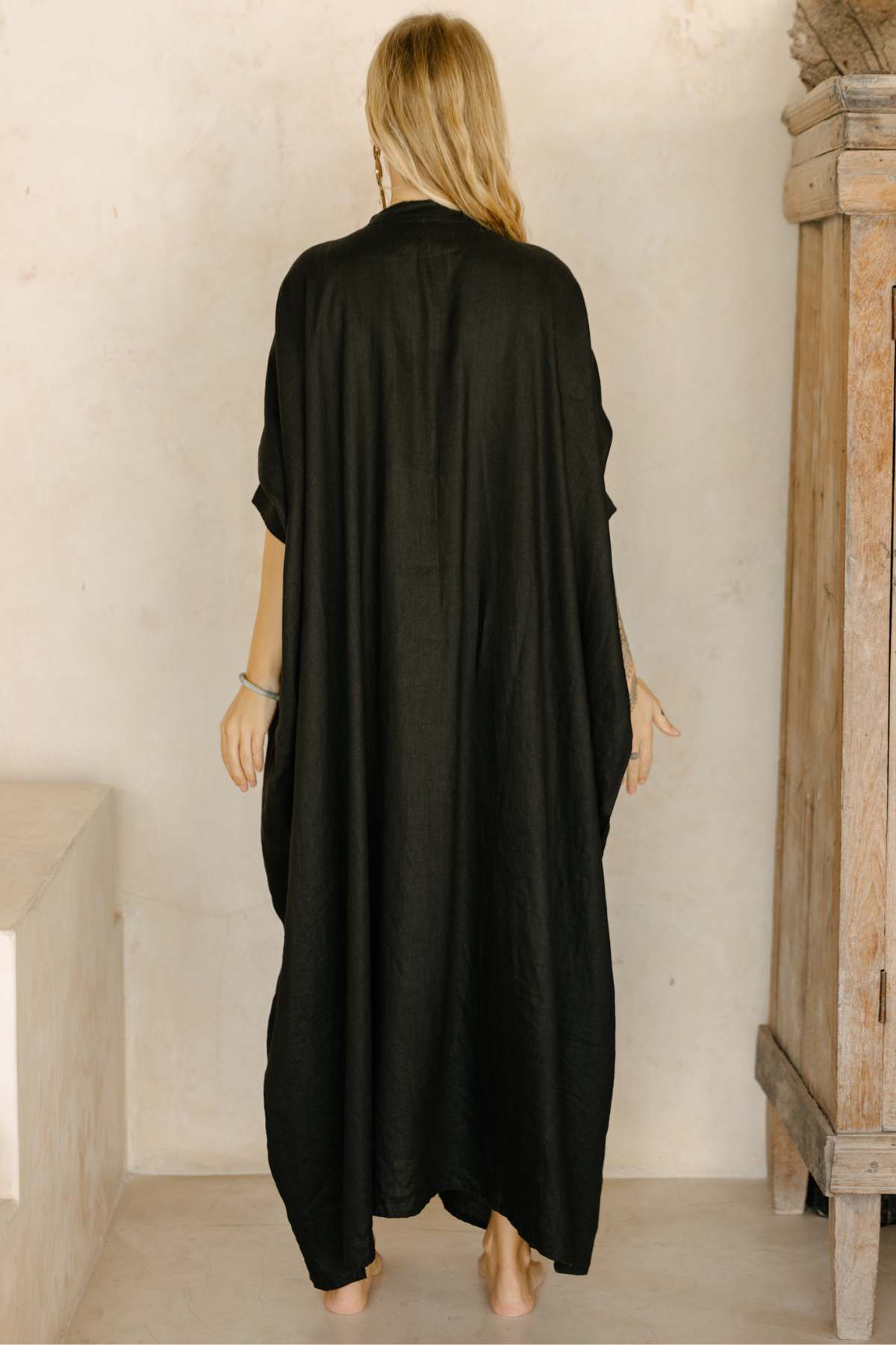 A woman with blonde hair, seen from behind, is wearing the La Majia Kaftan by Myrah Penaloza. The long, flowing black linen dress reaches her ankles as she stands in a minimalist room with light-colored walls and wooden furniture, evoking a sense of serene simplicity reminiscent of Frida Kahlo's artistic retreat.