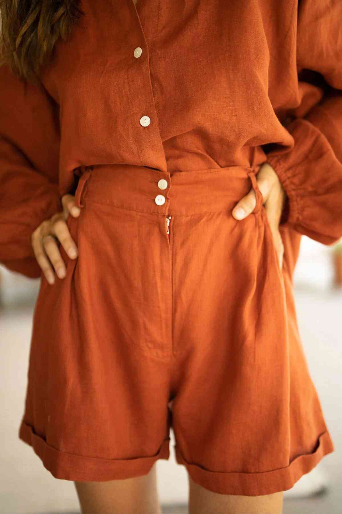 A person is wearing the elegant, rust-colored Rice Linen Set from Myrah Penaloza, crafted from stone-washed linen. The ensemble includes a long-sleeved, button-up top and high-waisted shorts that match perfectly. With their hands on their hips, they showcase versatile fashion centered around both the top and shorts.