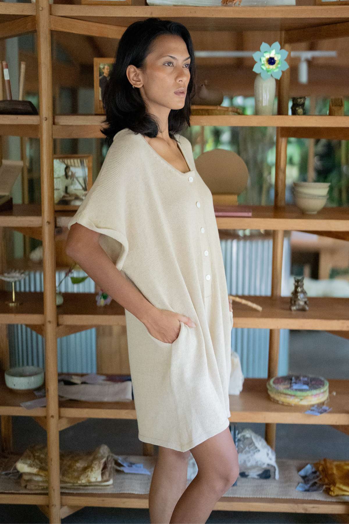 A woman in a Henley Knitted Playsuit - BF by Myrah Penaloza stands in front of wooden shelves filled with various decorative items. She looks to the side with a serene expression. The room has a cozy, eclectic ambiance, and her attire is subtly breastfeeding-friendly, adding an extra layer of comfort to the scene.