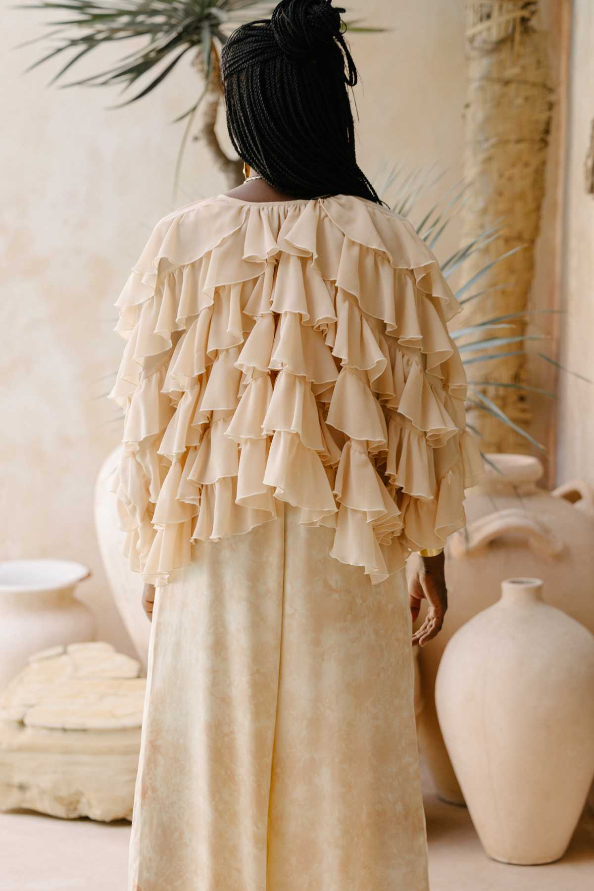 A person with braided hair stands with their back to the camera, showcasing the Anusara Bloom - Made To Order dress by Myrah Penaloza, featuring multiple layers of ruffles and a beige-colored floor-length skirt. The background includes large ceramic vases and a textured wall adorned with a plant, perfectly complementing the limited edition design.