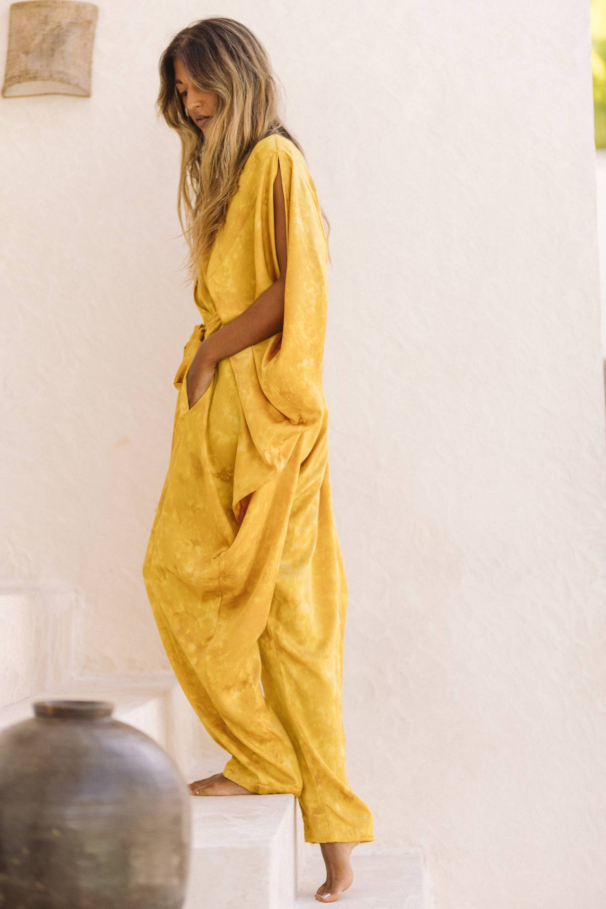 A person with long hair is descending white steps, wearing a loose, flowing Kuan Yin Playsuit in 100% silk and botanically dyed Turmeric Gold by Myrah Penaloza. The background consists of a plain, off-white wall and a large clay pot placed on one of the steps. The scene exudes a relaxed and serene atmosphere reminiscent of French linen charm.