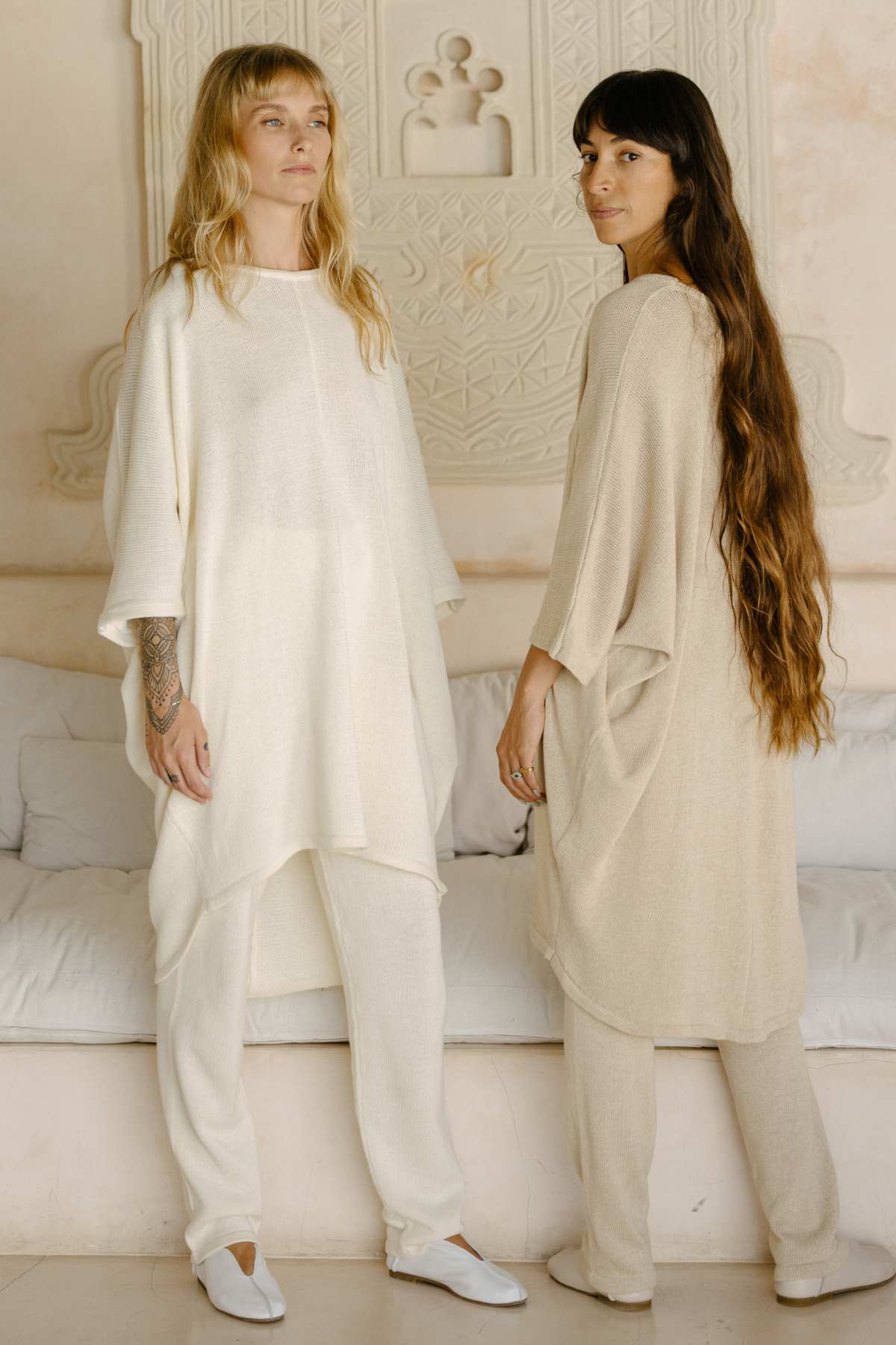 Two women stand indoors wearing loose, cream-colored outfits made from sustainable fabric. The woman on the left has shoulder-length blonde hair and is looking forward with her hands relaxed at her sides. The woman on the right, in a Butterfly Tunic Cotton Knit by Myrah Penaloza, has long dark hair, is turned slightly, and gazes towards the camera.