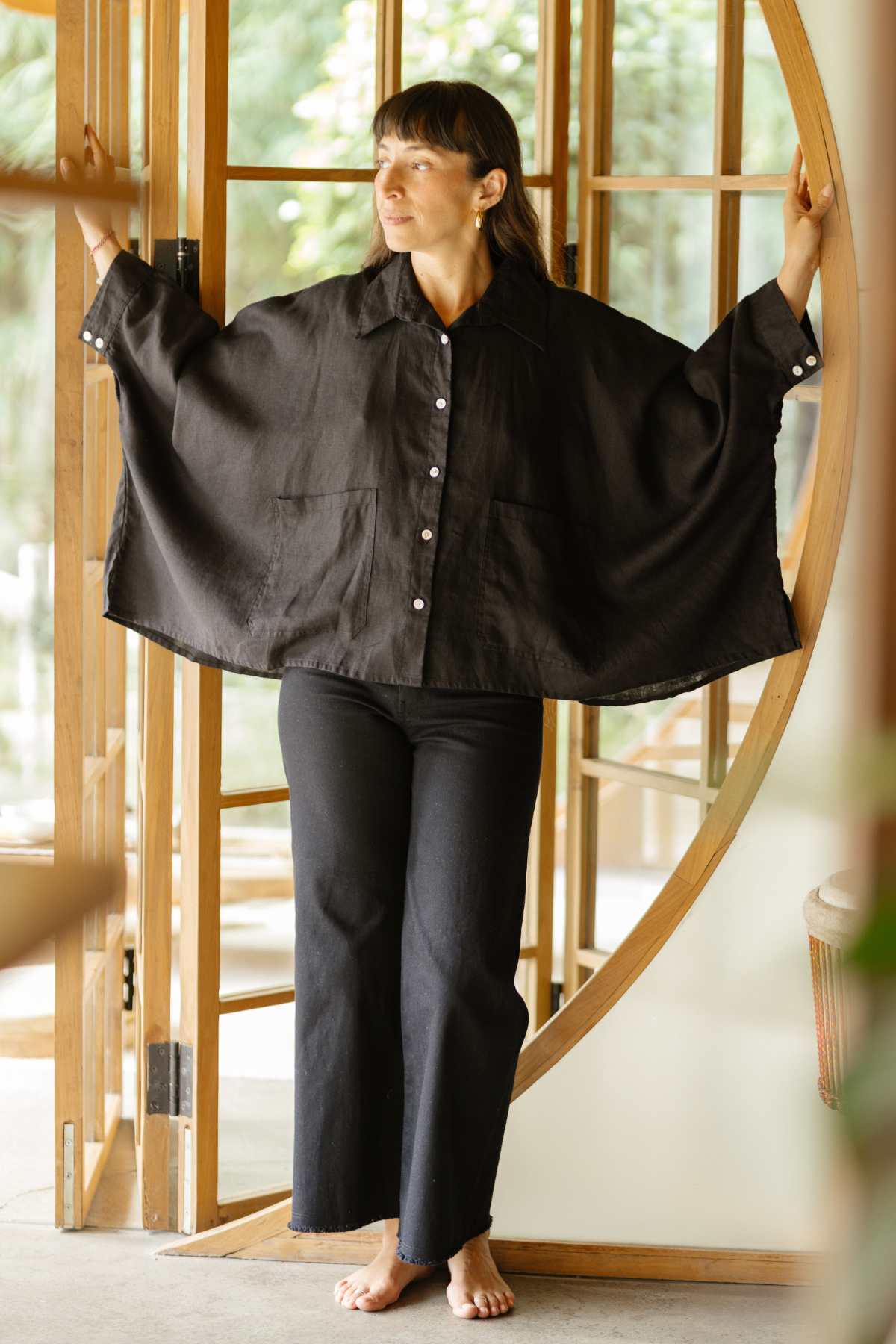 A person stands barefoot in a doorway with wooden framing, wearing the elegant Grace Linen Jacket by Myrah Penaloza paired with black pants. Sunlight filters through the glass, illuminating the peaceful indoor scene, perfectly complemented by the room's breathable design.