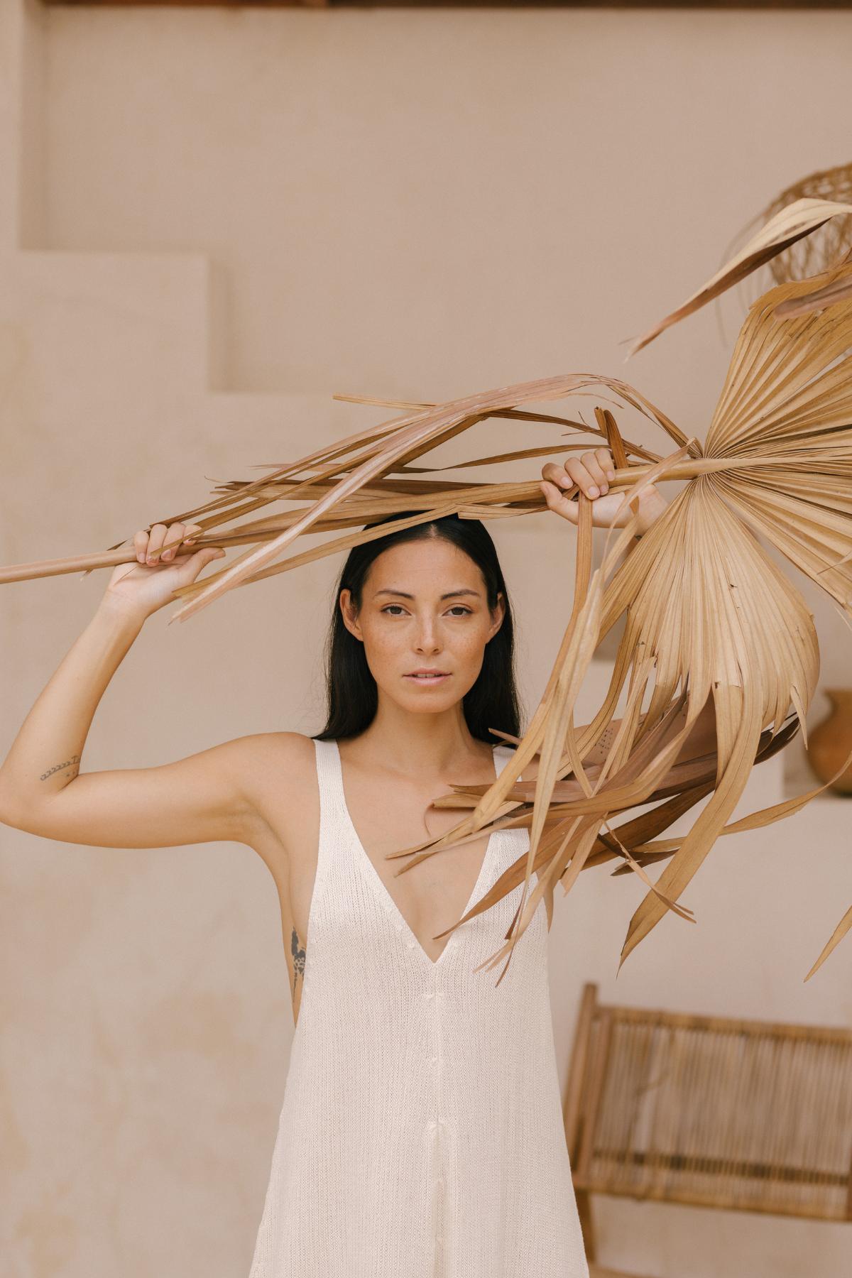 A woman with long, dark hair wears the Kundalini Playsuit by Myrah Penaloza in a white cotton knit, holding dry palm leaves above her head to highlight this versatile ensemble. She stands against a neutral-toned background with a wicker chair partially visible, exuding timeless elegance.