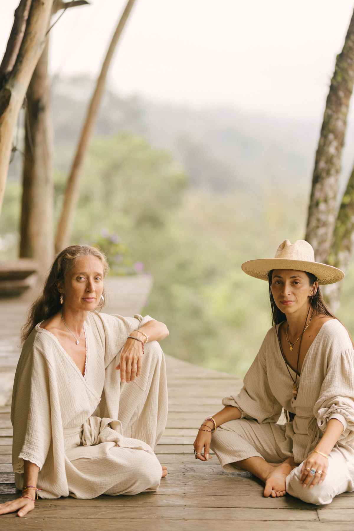 Two women in loose, light-colored clothing sit on a wooden deck outdoors, surrounded by lush greenery. One woman with long hair sits cross-legged wearing a Limited Edition Hand Embroidered River Playsuit by Myrah Penaloza, while the other woman with a wide-brimmed hat sits with her legs folded to the side.