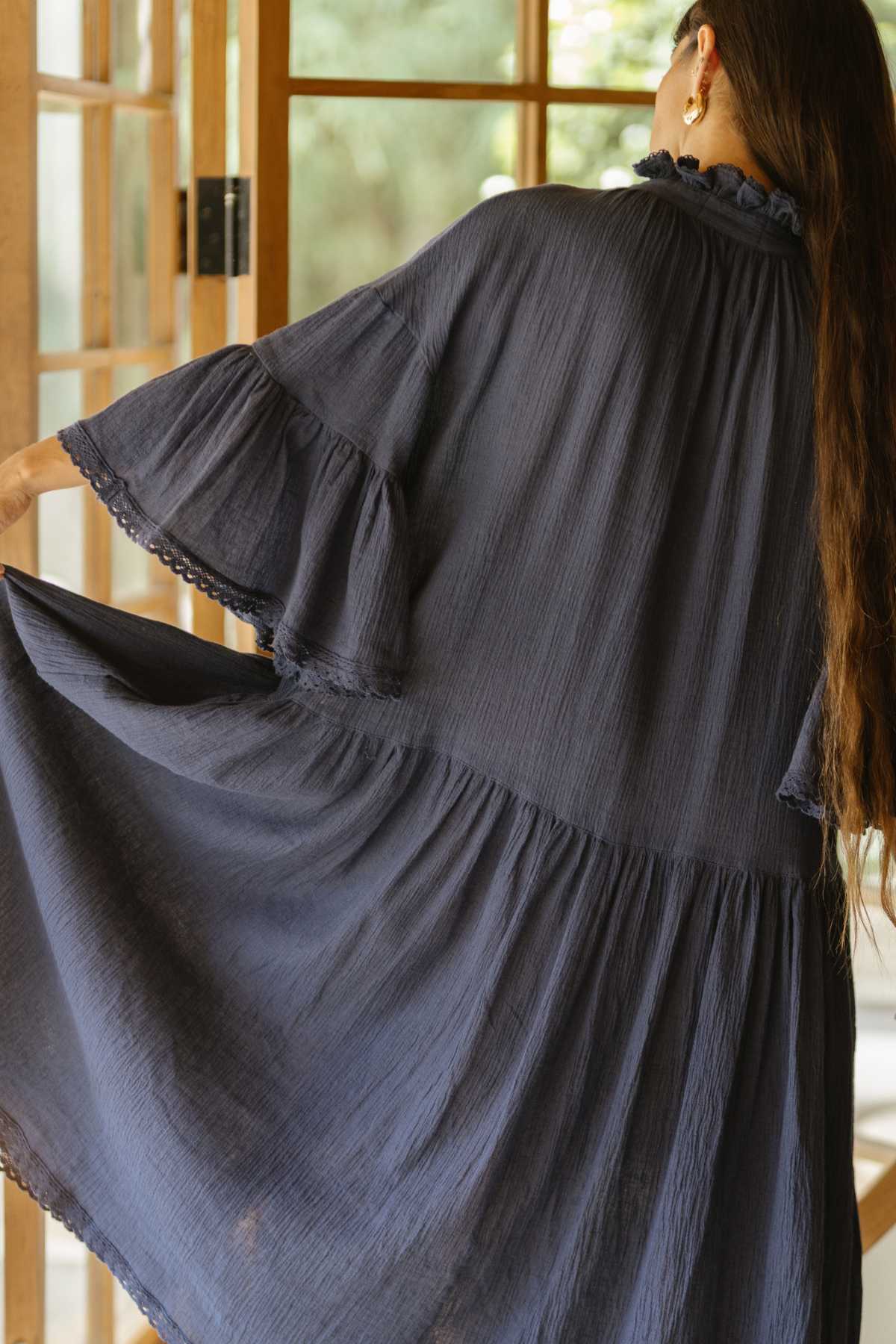 A person with long hair is shown from the back, wearing a flowing Myrah Penaloza gown called The Juliette Linen Gown in dark blue. They stand before a window with wooden frames, allowing natural light to filter through. The atmosphere is serene and elegant.