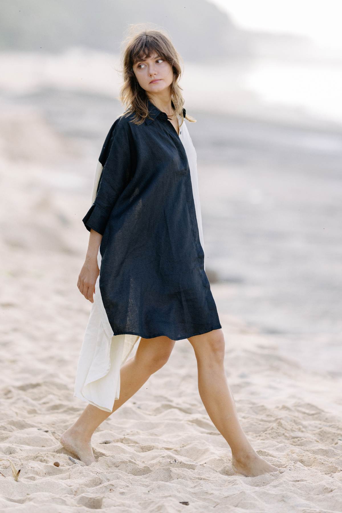 A woman with shoulder-length hair walks barefoot on a sandy beach, wearing a versatile Linen Suka Button Down Long Top by Myrah Penaloza. The background is blurred, featuring the shoreline and distant trees. The lighting suggests it's either morning or late afternoon.