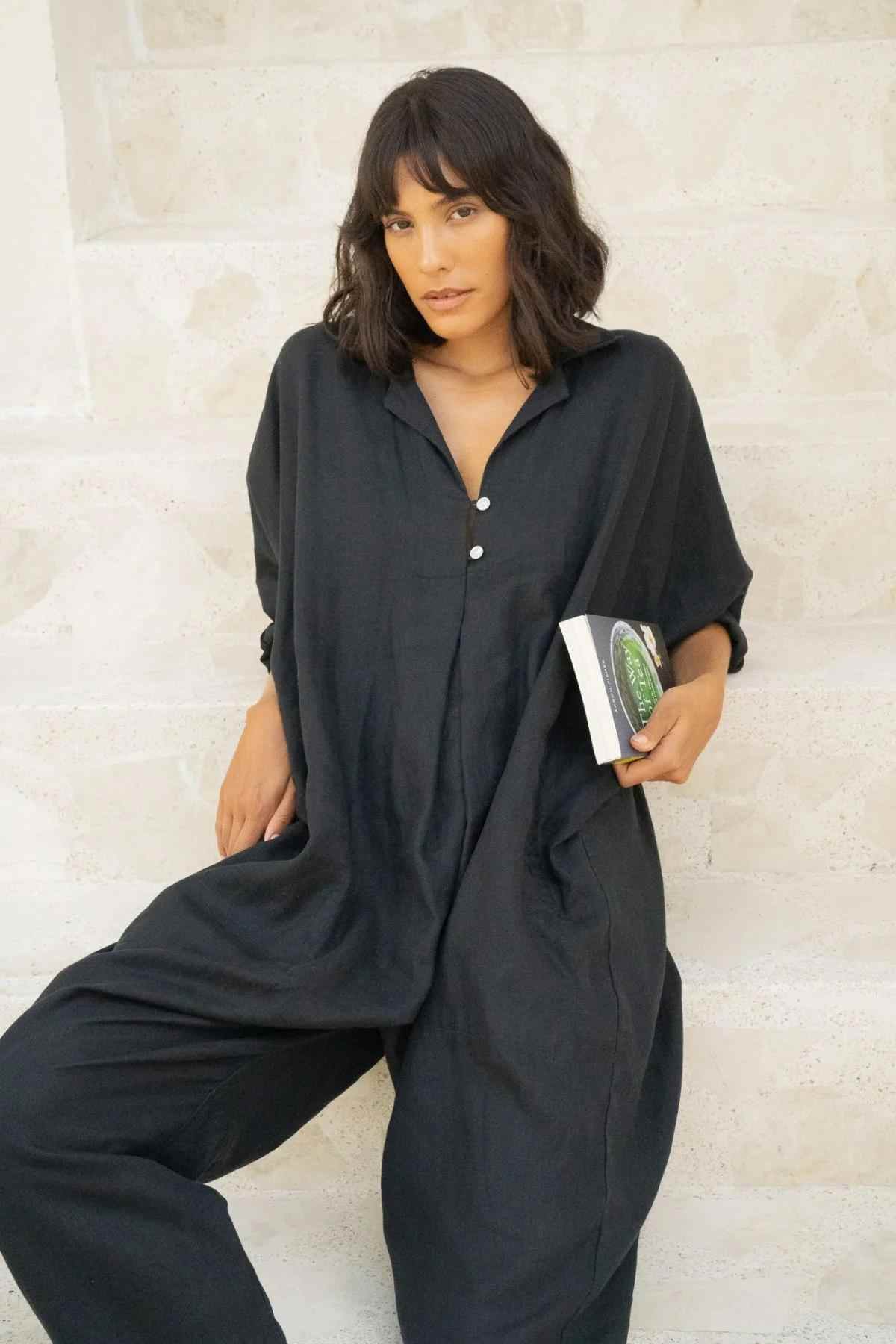 A person with shoulder-length dark hair is leaning against a rough-textured wall while wearing the Linen Sat Torri Playsuit by Myrah Penaloza, featuring a loose fit and wide dolman sleeves. They hold a small book with a partially visible green cover in one hand and gaze intently at the camera.