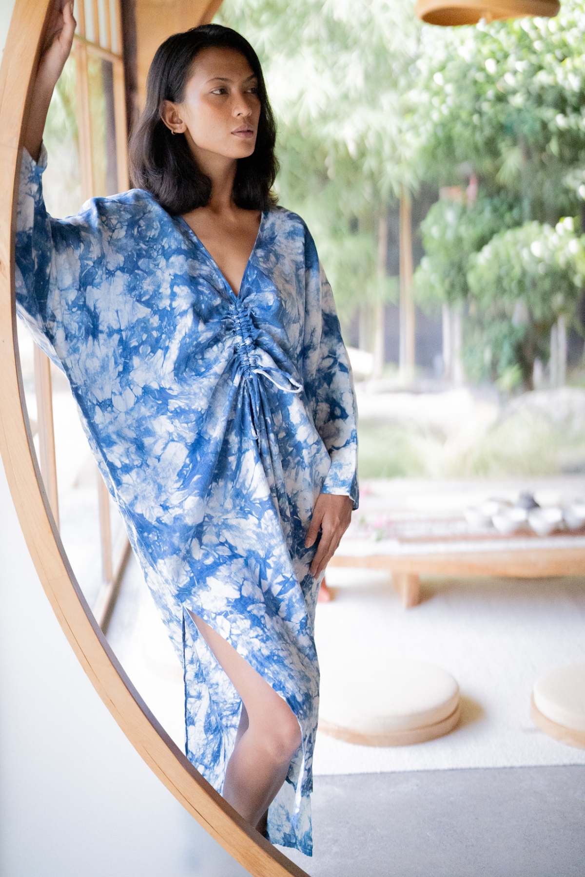 A woman wearing the Indigo Virgo Rising Kaftan by Myrah Penaloza stands near a circular wooden frame, gazing thoughtfully outside. The room showcases large windows with views of lush greenery and minimalist decor.