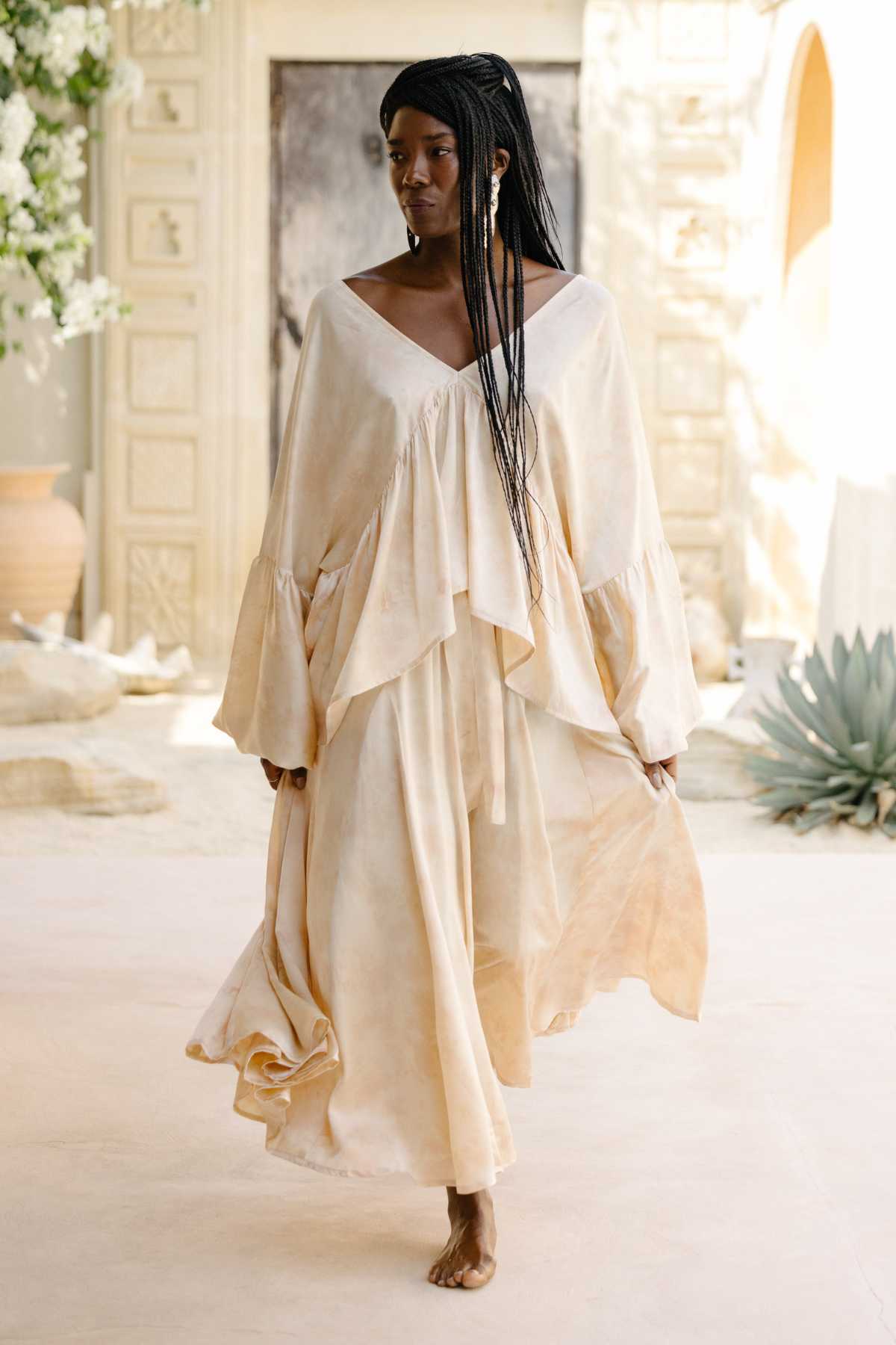 A woman with long braided hair walks barefoot in a flowing, botanically hand dyed silk ensemble from Myrah Penaloza. Her outfit consists of the Eva Top paired with the Orisa Dance Skirt, featuring ruffled sleeves and a cream-colored design. She is in a sunlit setting surrounded by large plants and decorative stone walls.