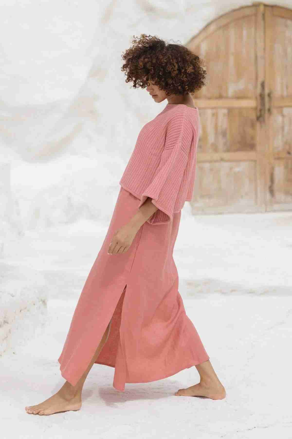 A person with curly hair is walking barefoot on a white surface, dressed in the Spirit Top Sweater Knit by Myrah Penaloza. The ensemble consists of an oversized pink top paired with a long skirt featuring side slits, all made from luxurious cotton fabric. In the background, a large wooden door is visible.