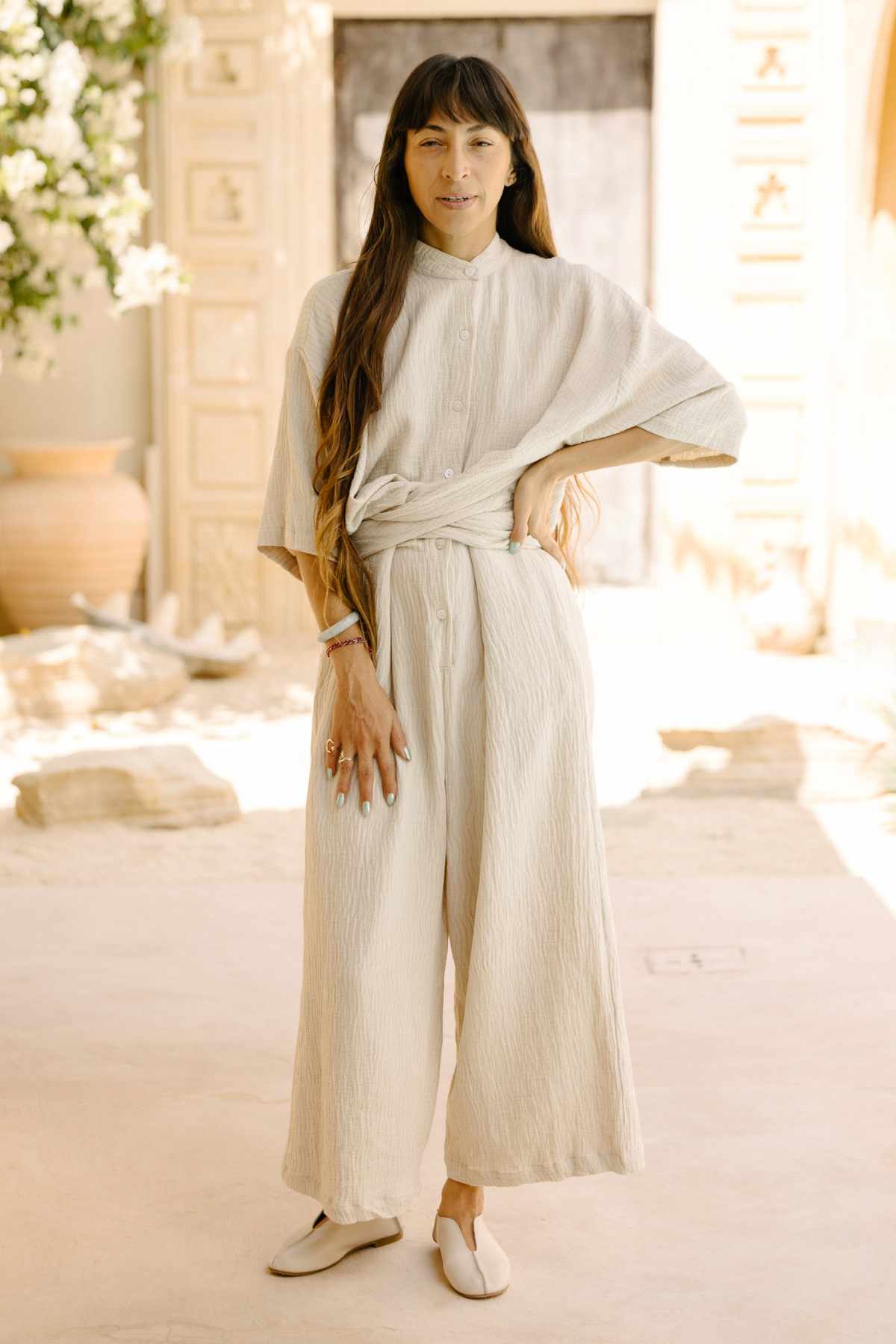 A person with long hair stands in a light, airy room wearing the cream-colored Pine Playsuit, a cotton/linen blend by Myrah Penaloza. They are barefoot, and the background includes a large vase, a wooden door, and soft, natural lighting.