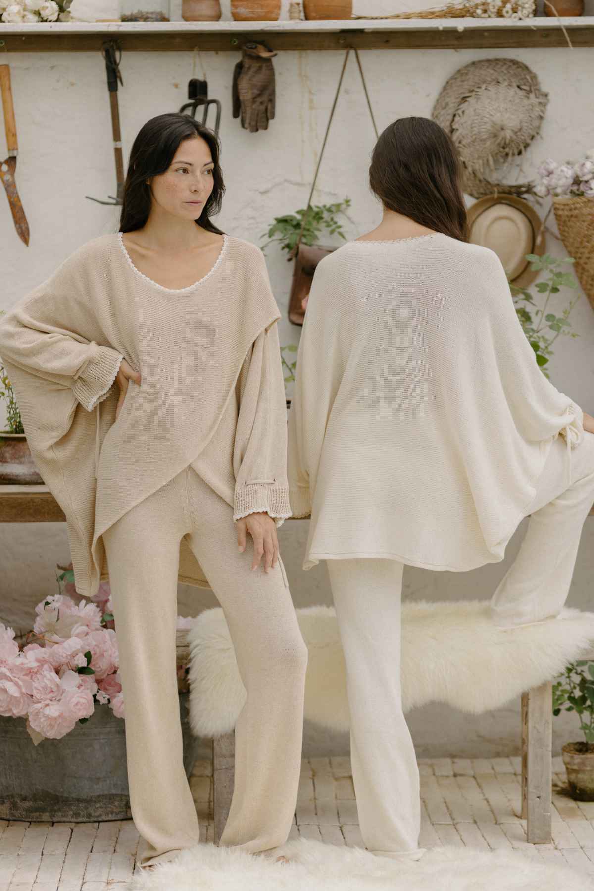 Two women are modeling cozy, neutral-toned loungewear. The woman on the left faces forward, wearing a beige Honey Knit Sweater and matching pants from Myrah Penaloza. The woman on the right, seen from behind, wears a loose-fitting white top and matching pants. The decor includes baskets and flowers.