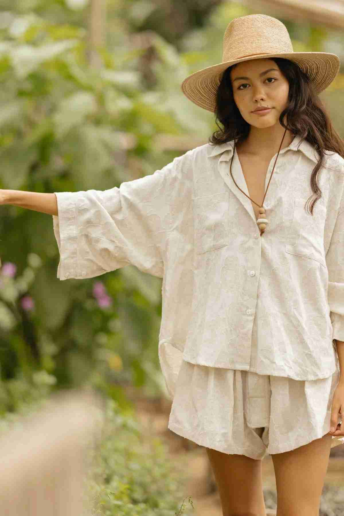 A woman in light, loose-fitting clothing and a straw hat stands in a garden, wearing the Linen Nidra Button Down Set by Myrah Penaloza. She pairs it with shorts as her hand rests on the wooden railing, surrounded by the lush green backdrop.