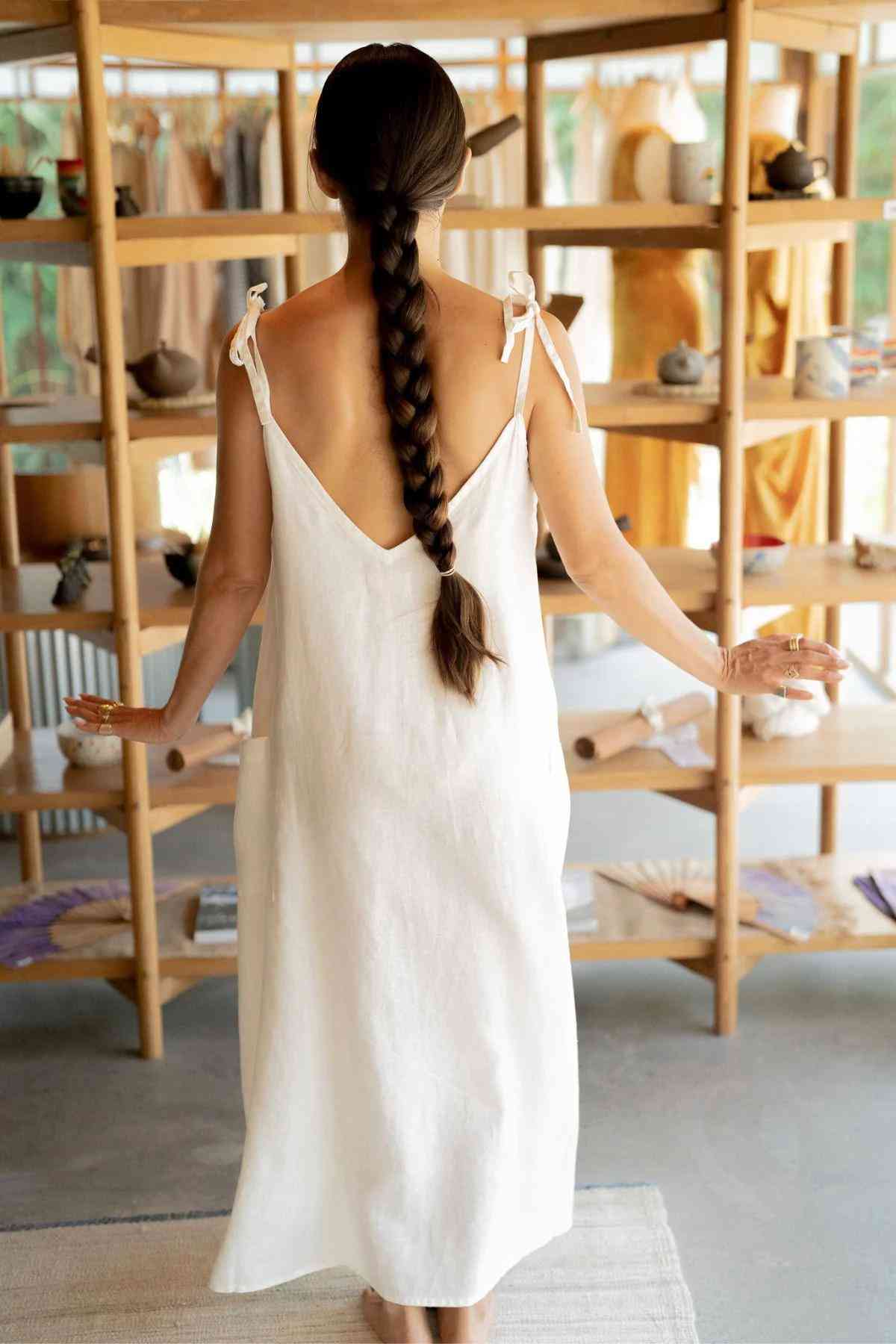 A person with a long braid, wearing a Radiant Gown - Made To Order from Myrah Penaloza, stands facing away in front of wooden shelves filled with assorted items. They have their arms slightly outstretched, creating a serene and calm atmosphere. Natural light filters into the room.