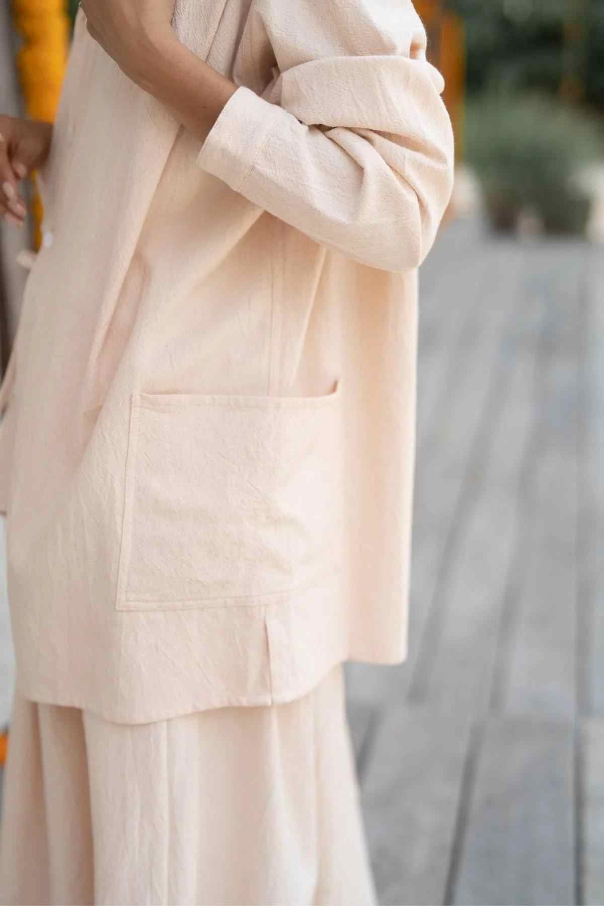 A person wearing the Myrah Penaloza Tender Jacket & Pant Set (3 of a kind) - Pre Order Only in beige stands outdoors on a wooden deck. The image focuses on the upper body and side, highlighting the long sleeve and front pocket of the jacket. A blurry background with greenery is visible.