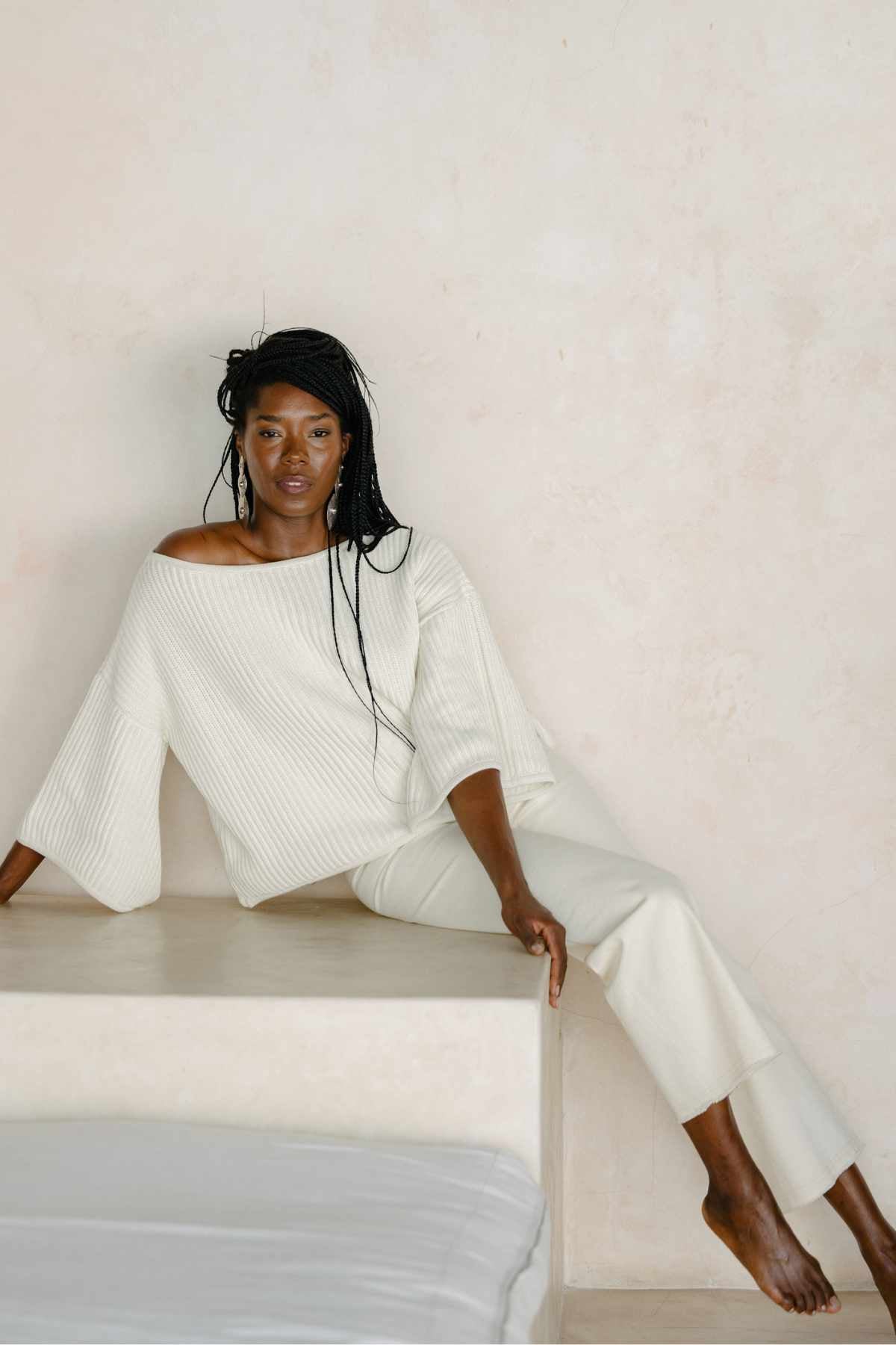 A woman with long braided hair sits on a ledge against a light-colored wall, dressed in the Spirit Top Sweater Knit from Myrah Penaloza and matching pants. The loose, off-shoulder, oversized top made from luxurious cotton fabric complements her calm expression as she poses barefoot with one leg bent, exuding a relaxed and elegant vibe.