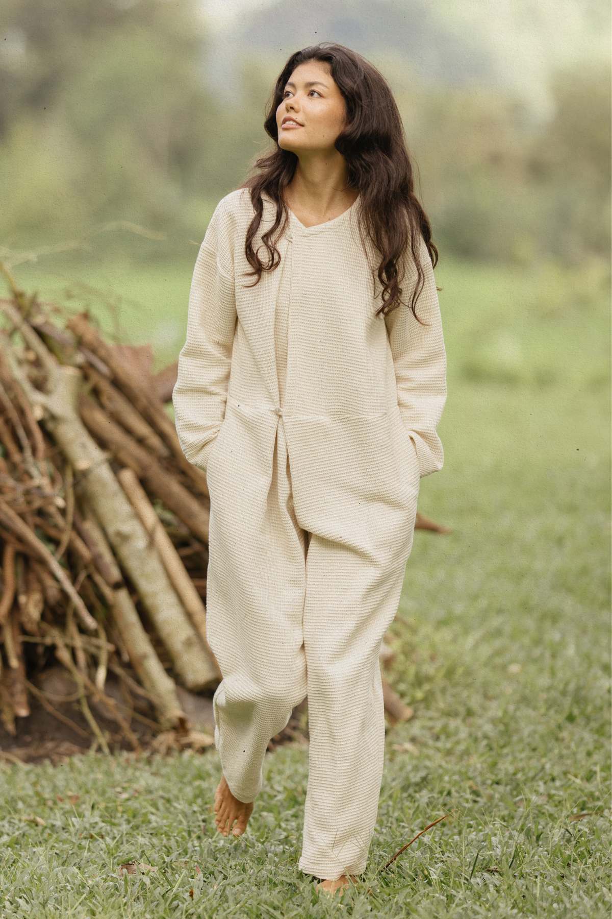 A person with long dark hair, wearing a loose, light-colored Camper Playsuit by Myrah Penaloza made of Waffle cotton, walks barefoot on a grassy area. A stack of logs and trees adorn the background as they look slightly upwards. The setting exudes the charm of adventurous weekend getaways in a natural environment.